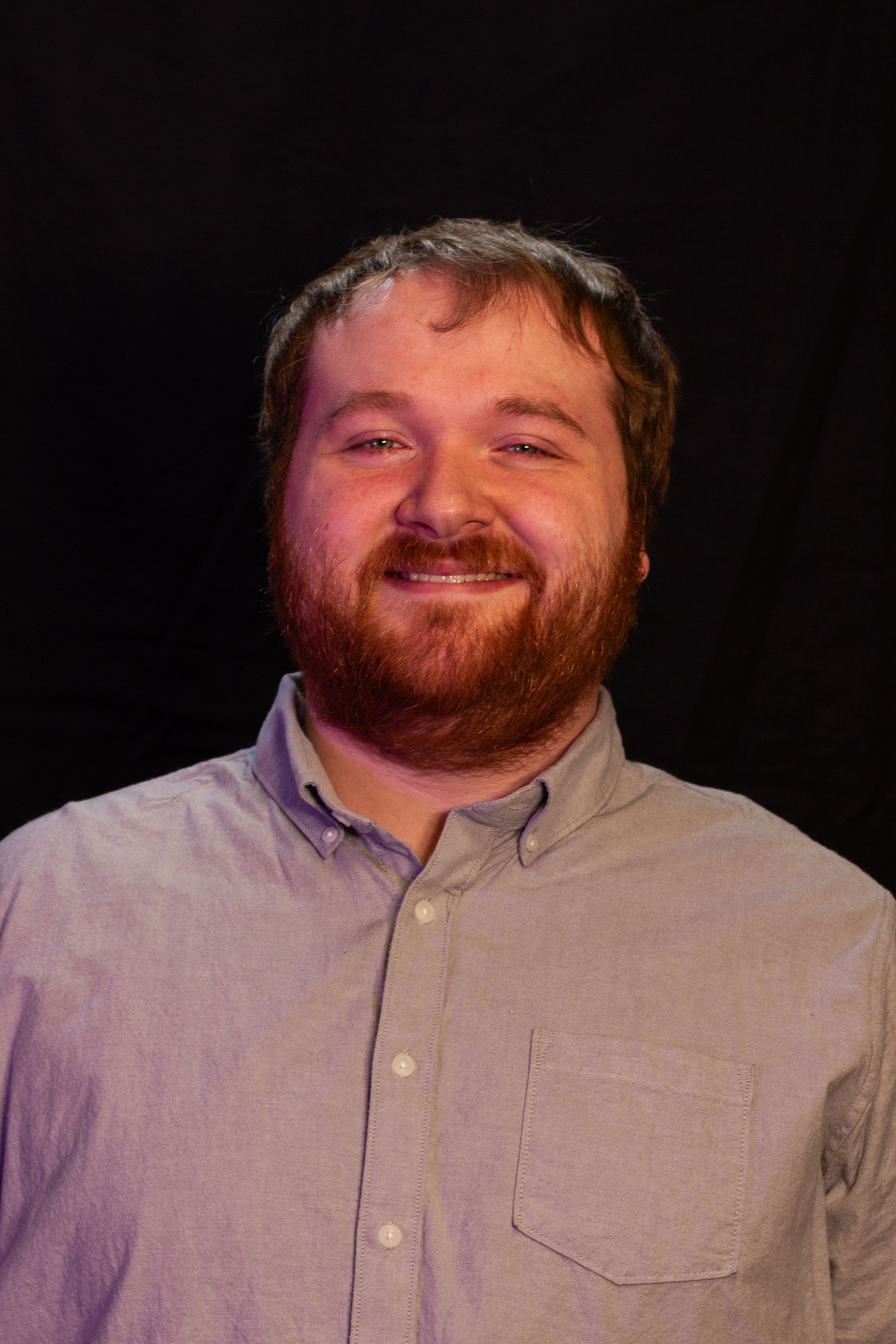 Man with beard, wearing button-down shirt, smiles in front of a dark background.