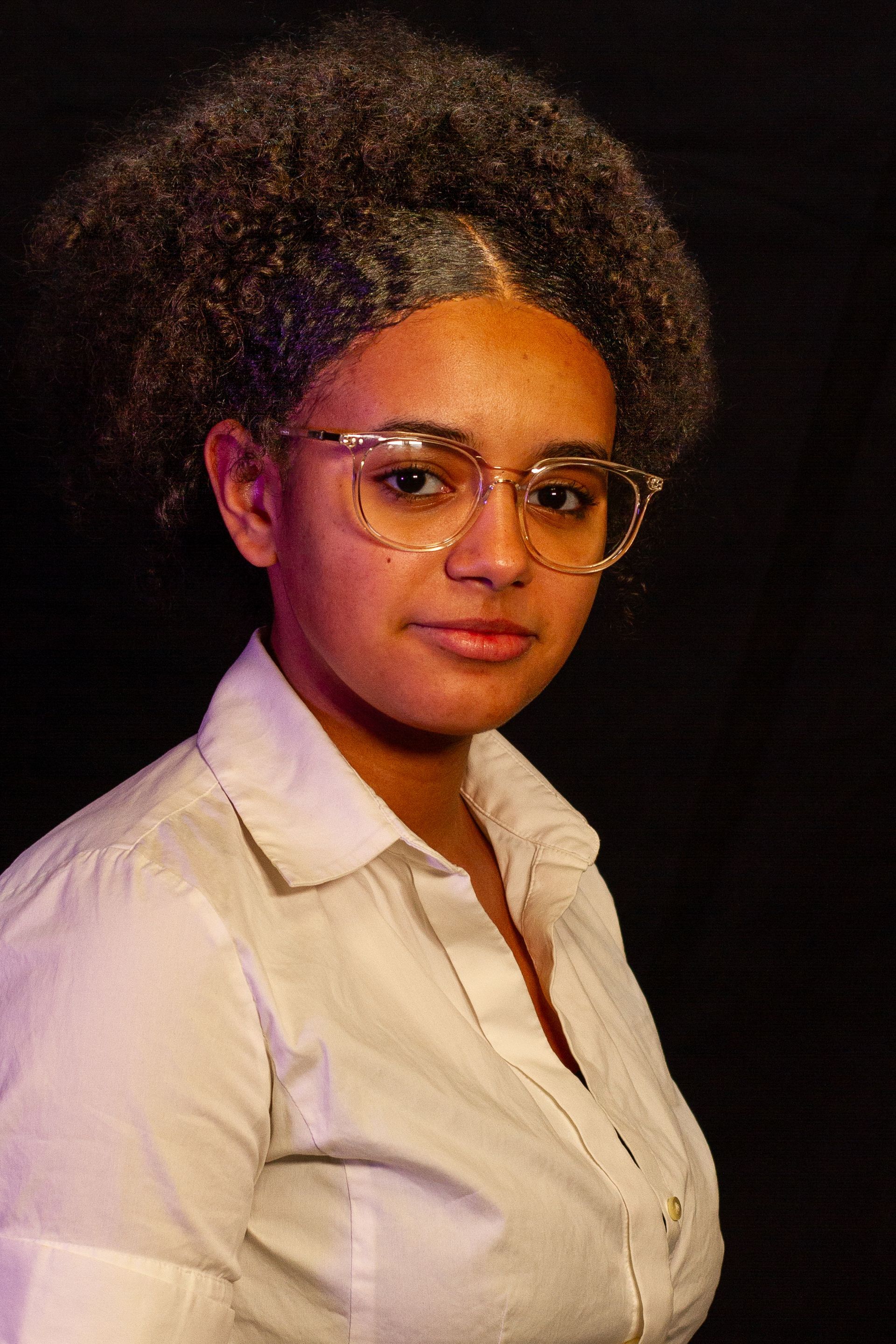 Woman with curly hair and glasses, wearing a white shirt, looking directly at the camera against a dark backdrop.