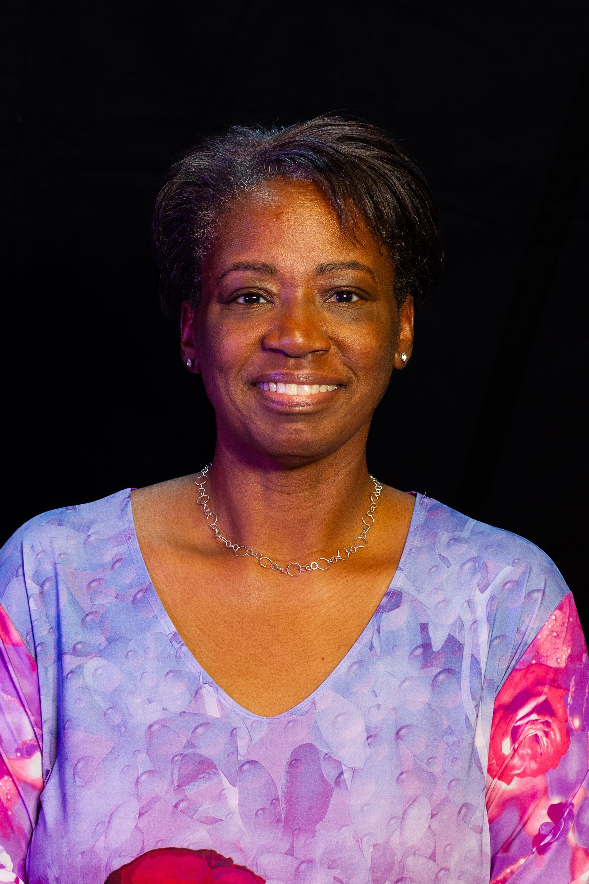Smiling Black woman with short hair wearing a floral blouse and silver necklace against a black background.