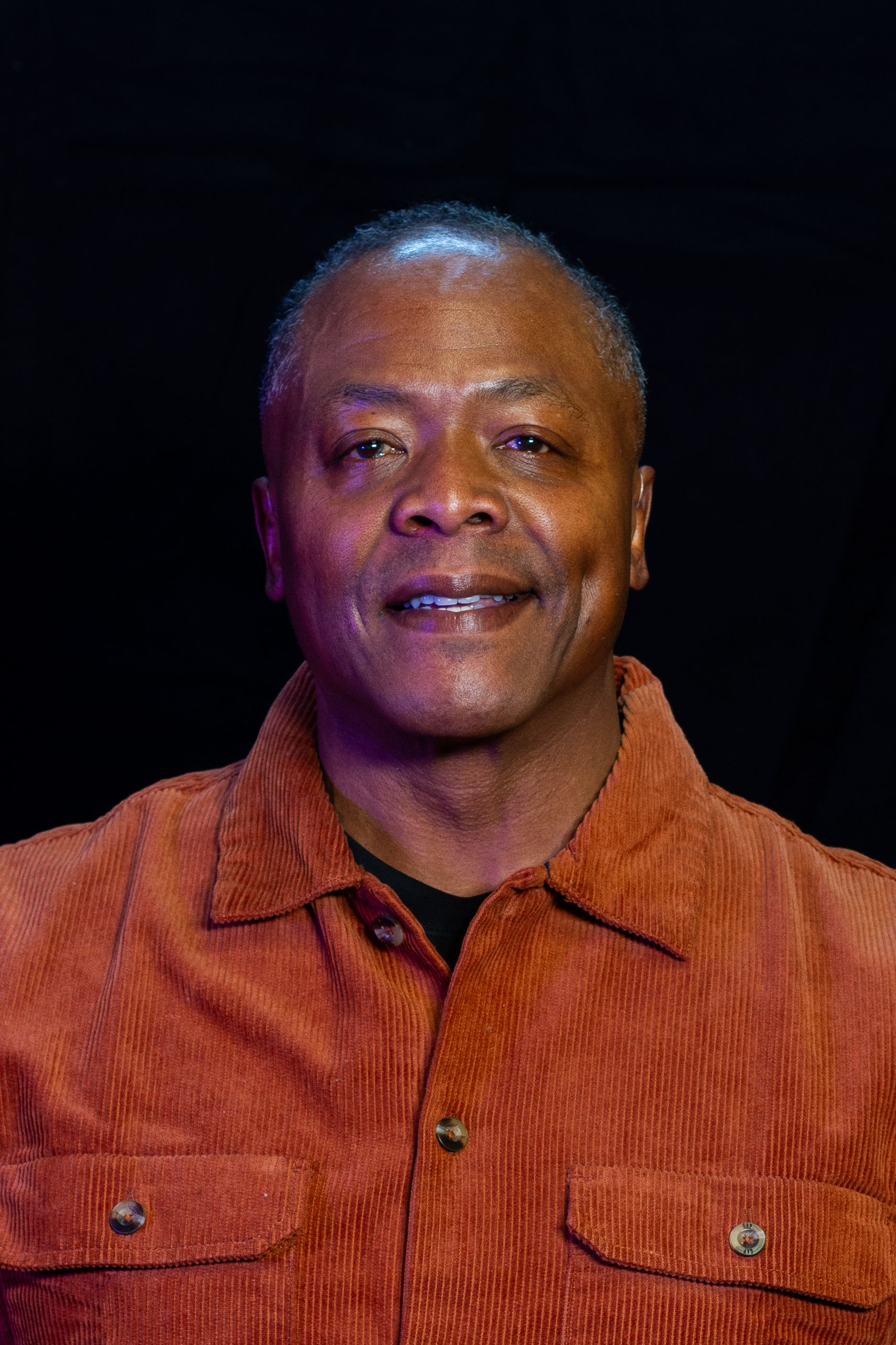 Mature Black man smiles, wearing orange corduroy shirt against a black background.
