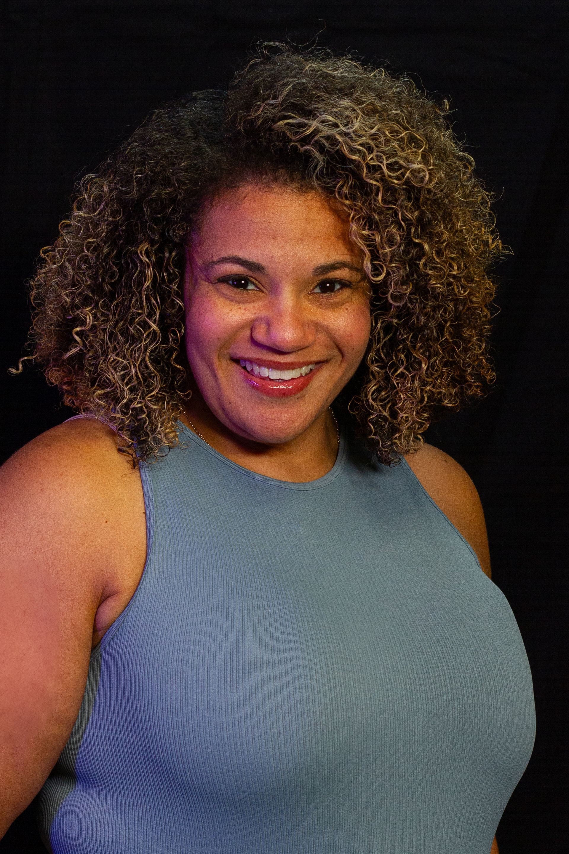 Woman with curly brown hair smiles, wearing a blue sleeveless top, against a black background.