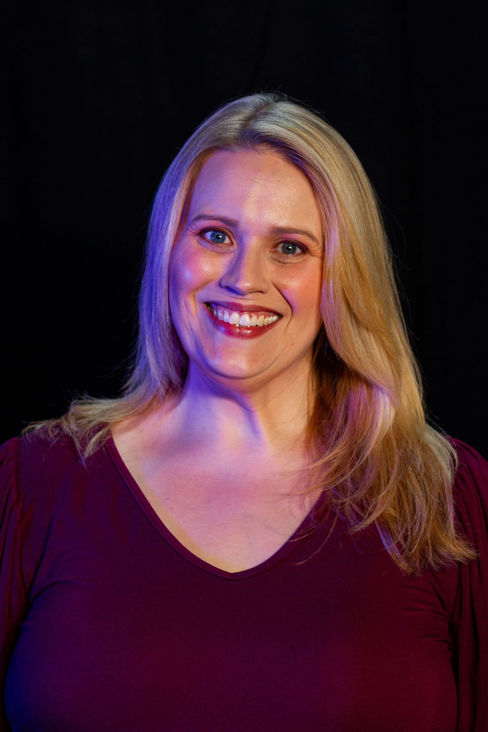 Woman with blonde hair smiles, wearing a burgundy shirt, against a black background.