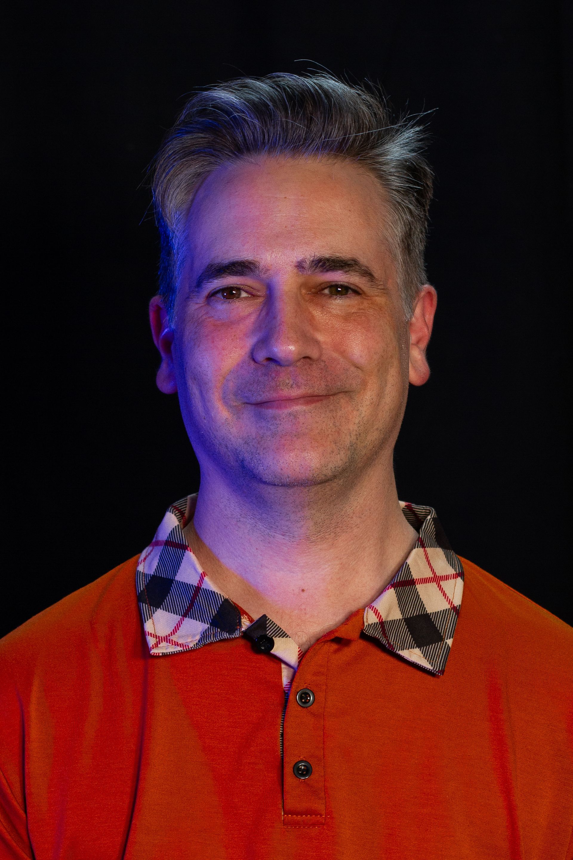 Man with grey hair, smiling, wearing orange shirt with plaid collar, black background.