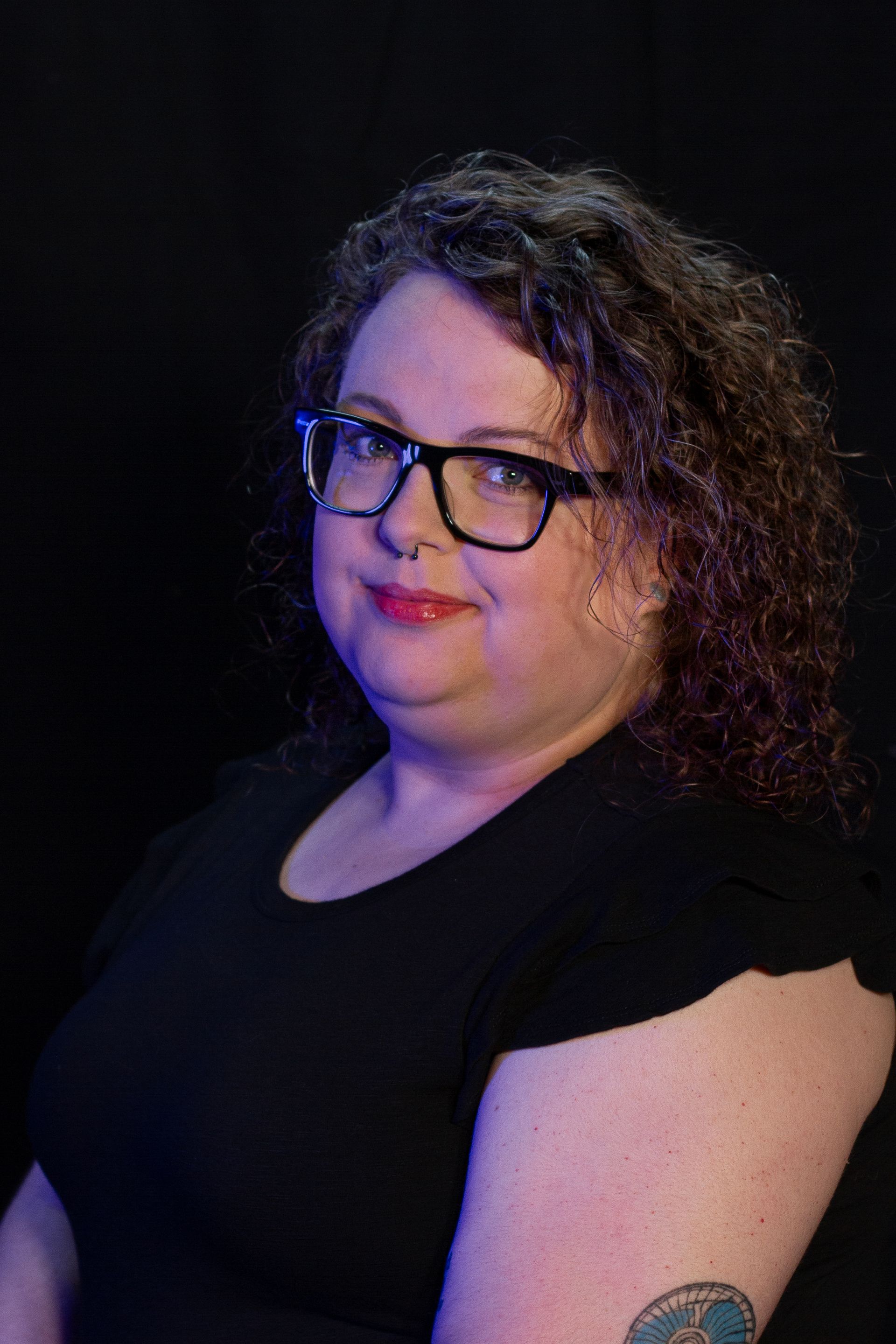 Woman with glasses and curly hair smiles, black top, dark background.