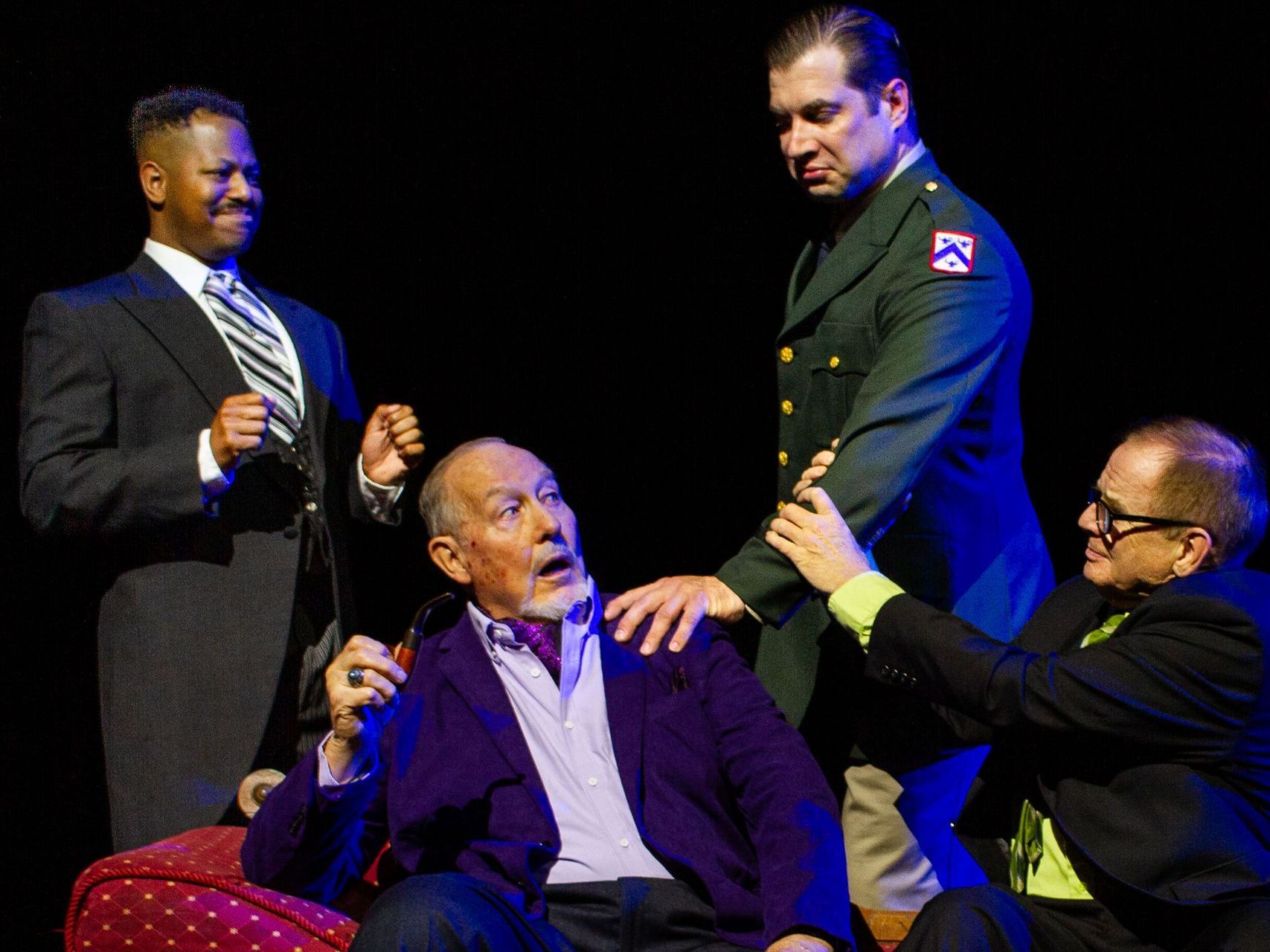 Four men in a dramatic scene: one seated, grabbed by others in a dark setting; one in a suit, one in a military uniform.