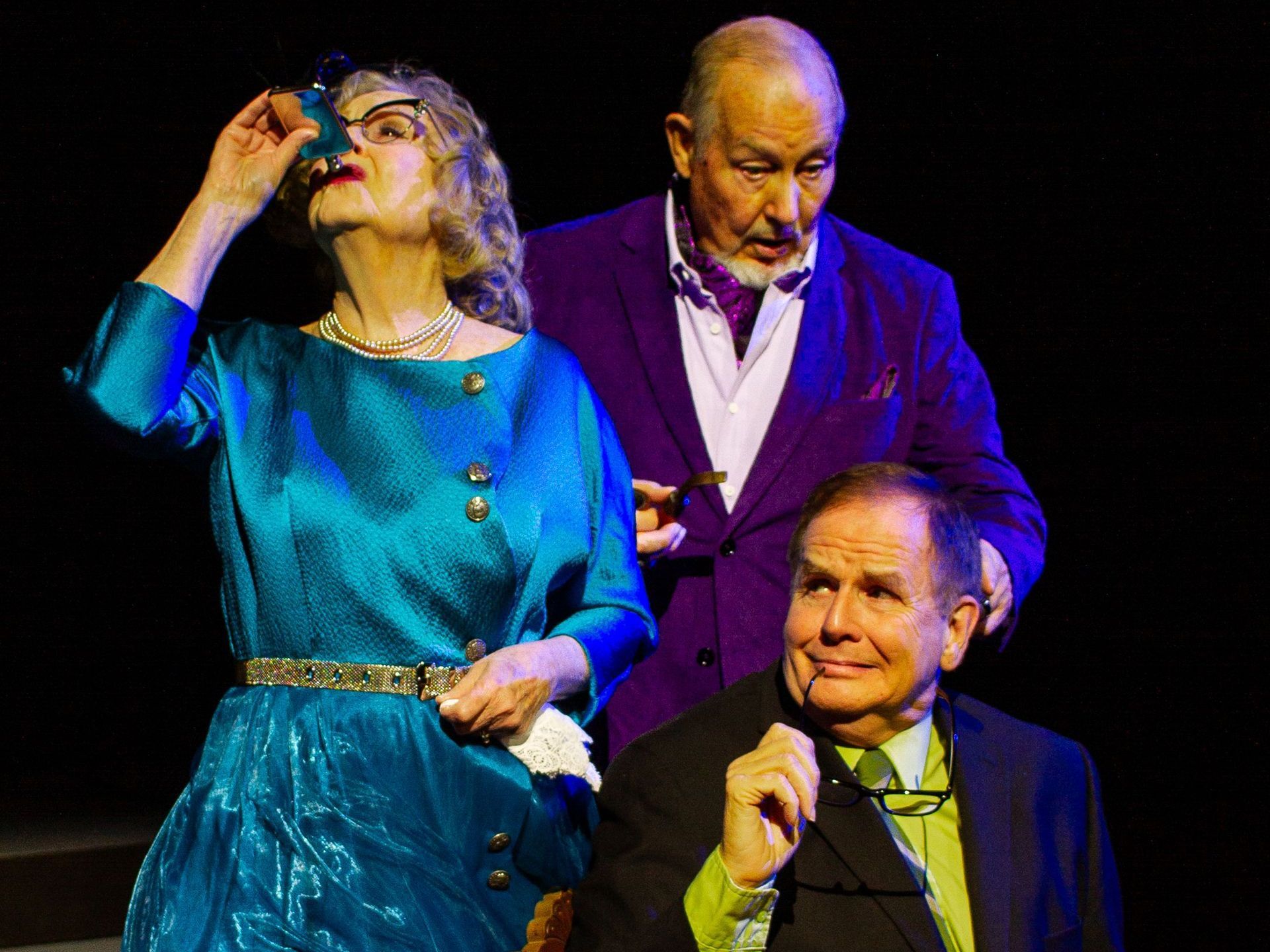 Three actors in a play: woman in blue looking up, man in purple, and seated man in a suit, all smiling.