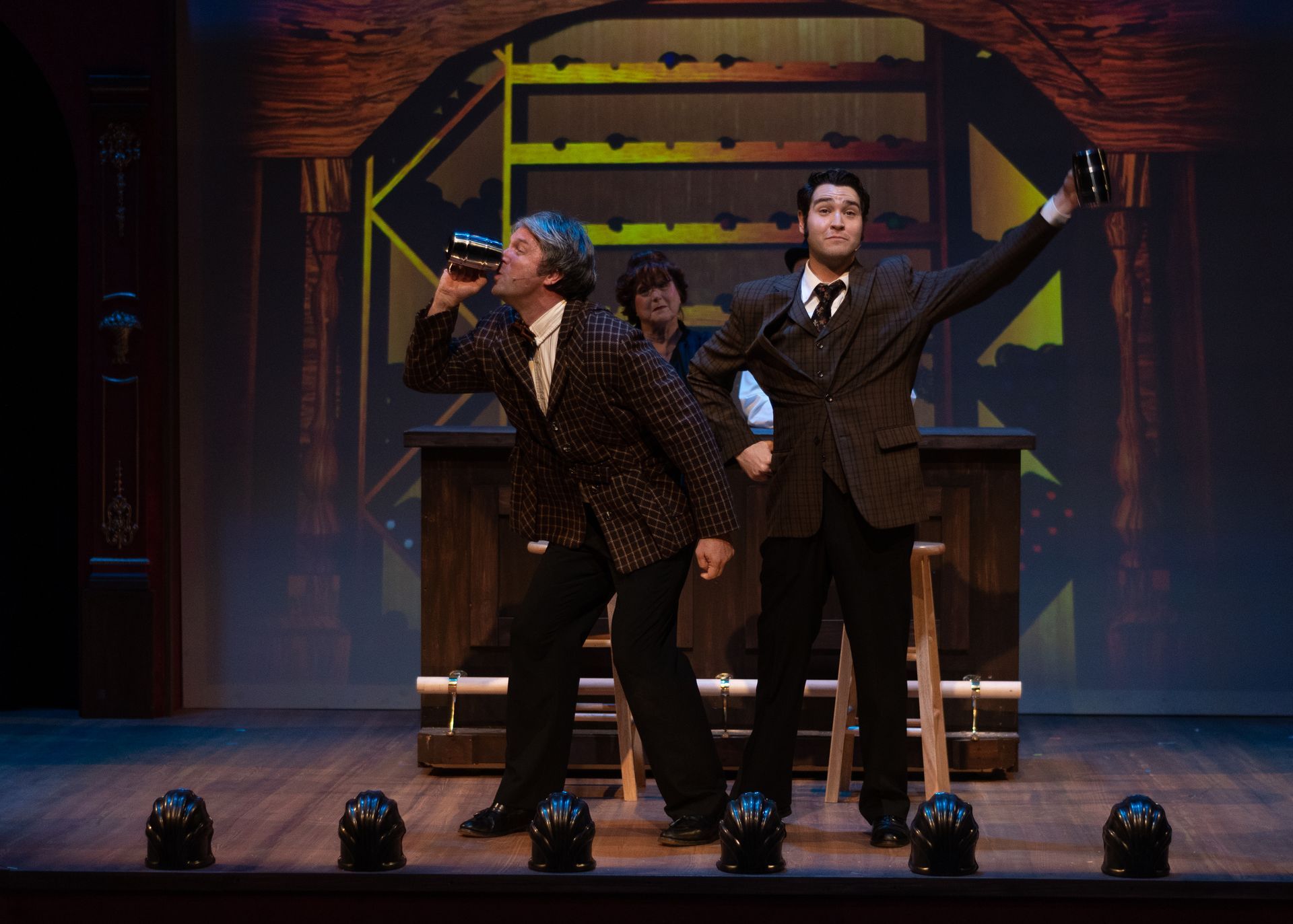 Men on stage at a bar, one drinking, the other raising a glass, backdrop of wine bottles.