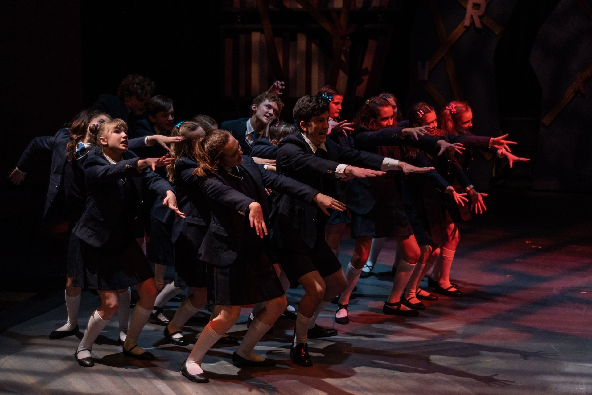 A group of schoolgirls in navy uniforms dance on a stage, arms outstretched.