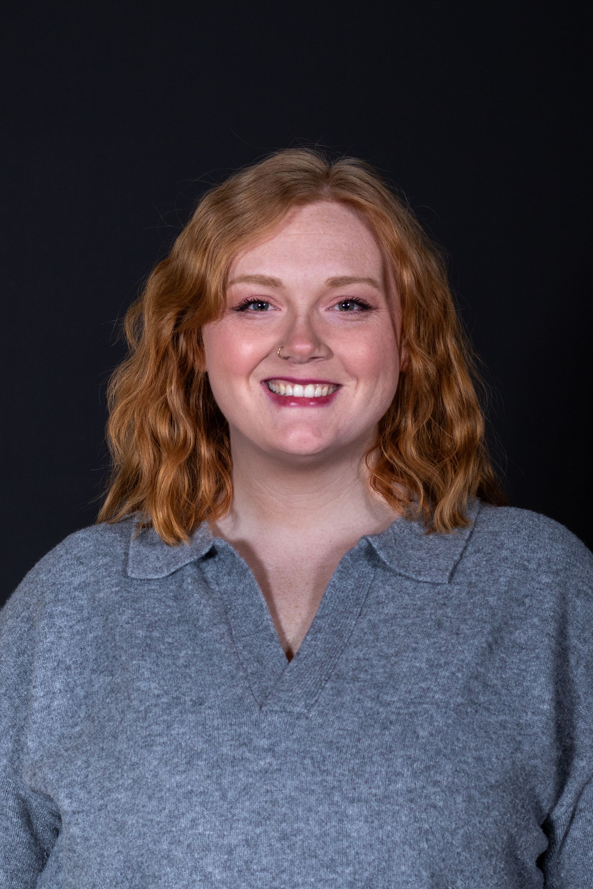 Woman with curly red hair, smiling in a grey sweater against a dark background.