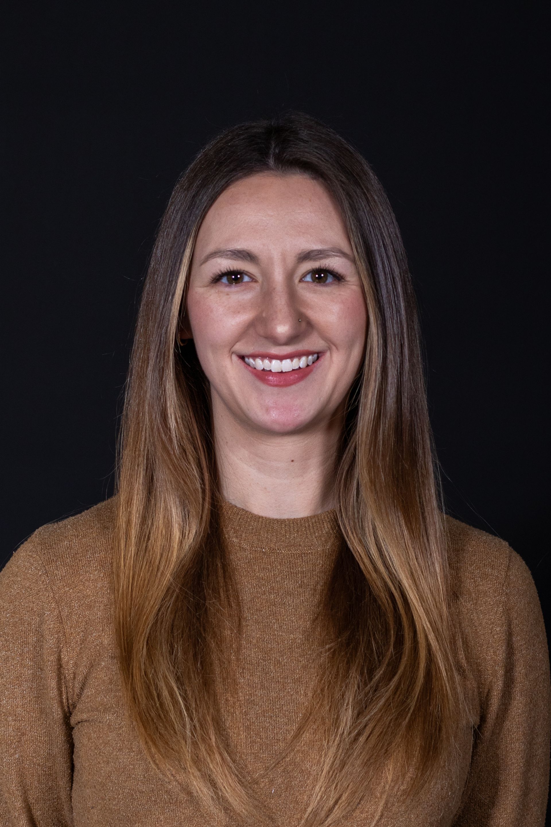 Woman with long, ombre hair smiles at the camera, wearing a gold-toned sweater, against a black backdrop.