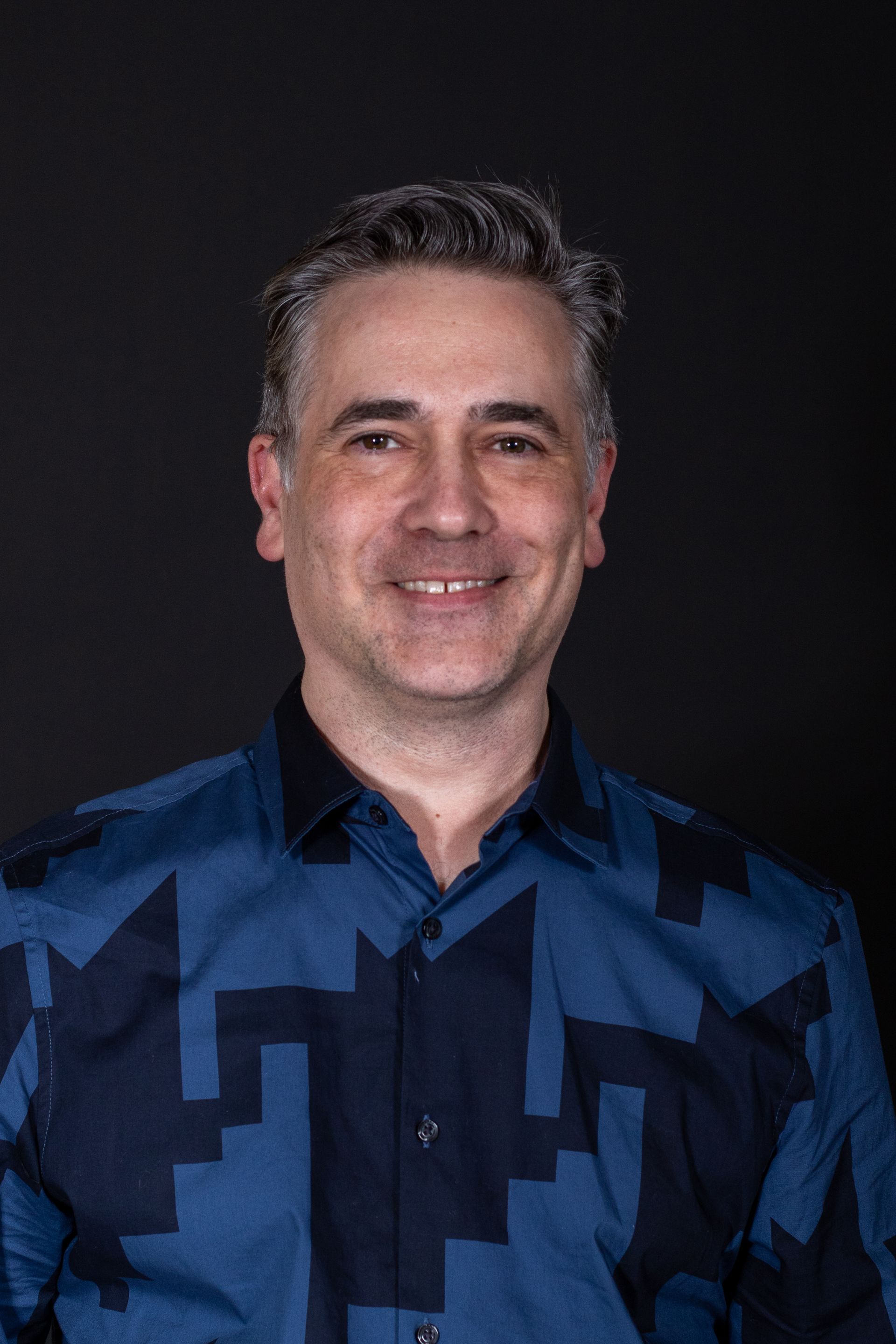 Man in blue patterned shirt smiling against a dark background.