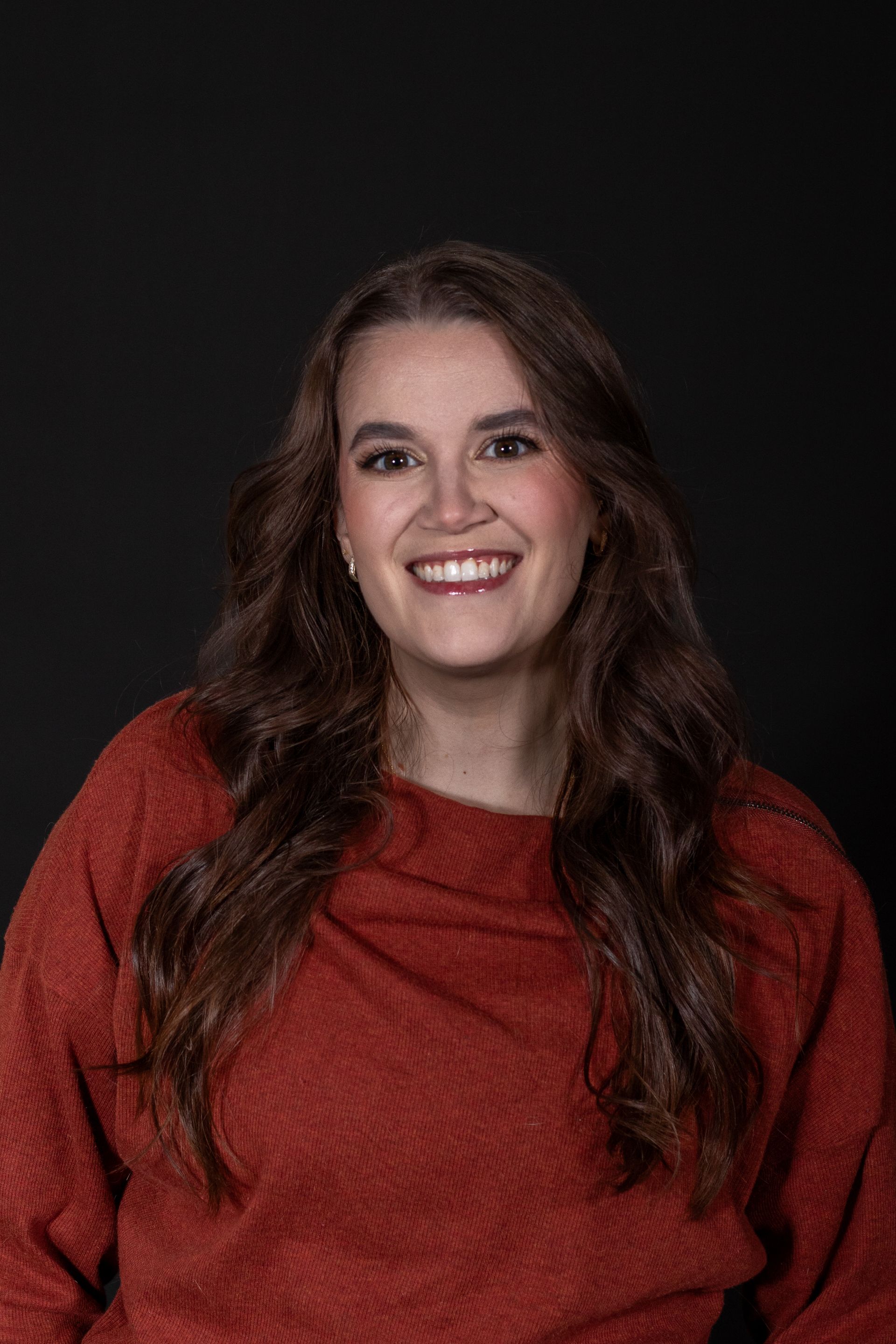 Woman with long brown hair smiling, wearing a rust-colored sweater against a black background.