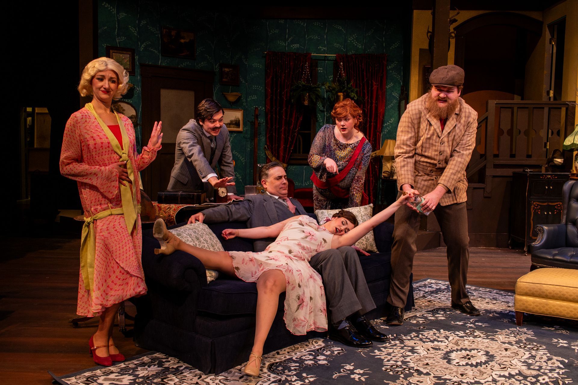 A play scene: Six people in vintage attire, some with surprised expressions, posed on a set with a blue couch and rug.