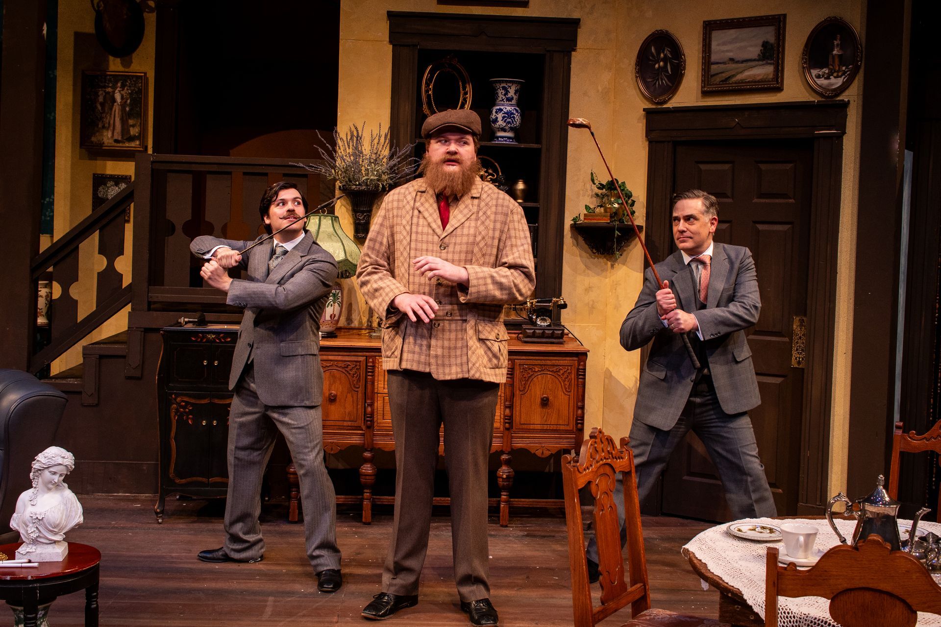 Three men on stage in a period play, posing dramatically. Interior setting with antique furniture.
