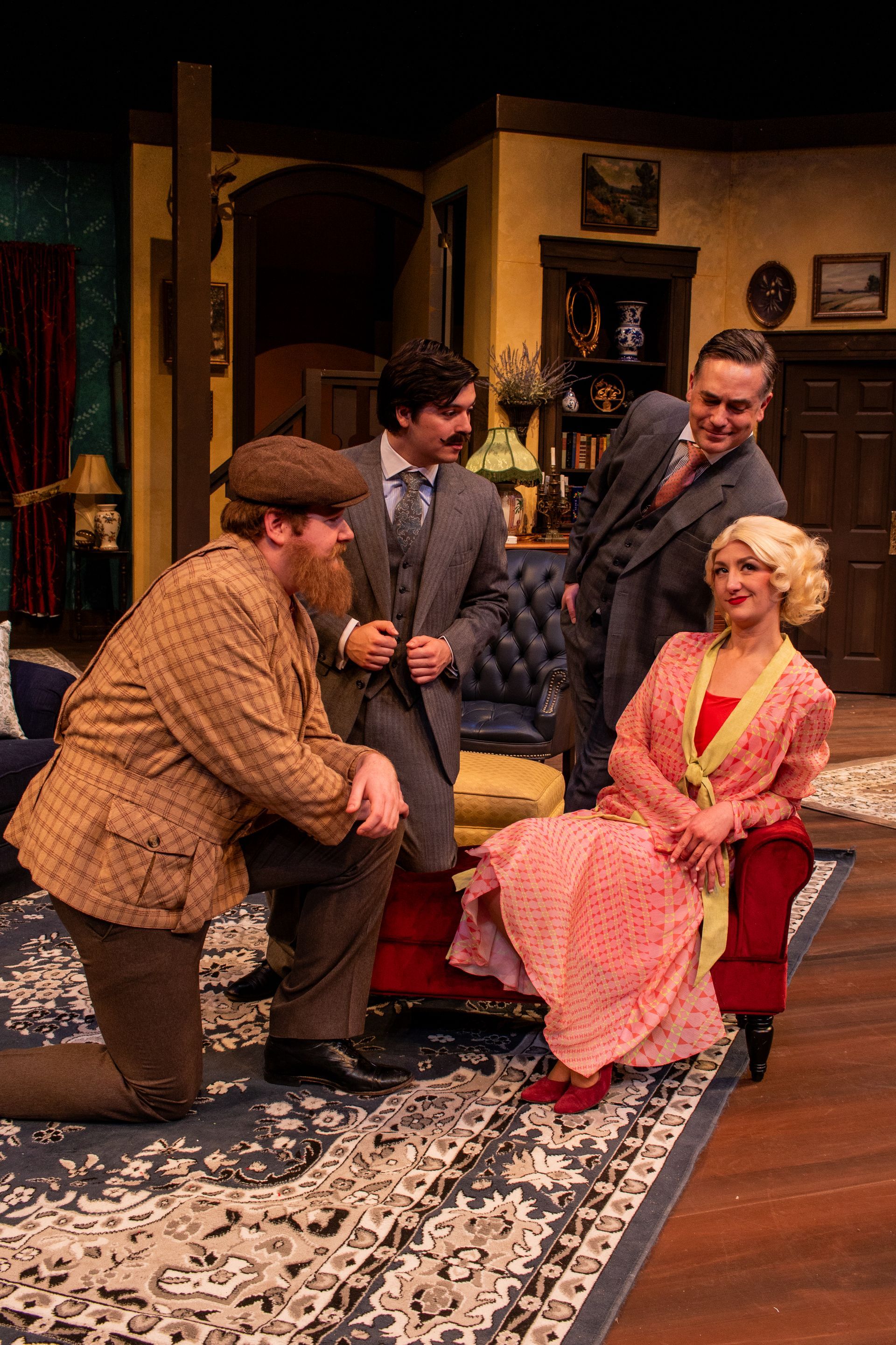 Four people in a room, dressed in period attire. Man kneels; others stand near a woman seated in a chair.