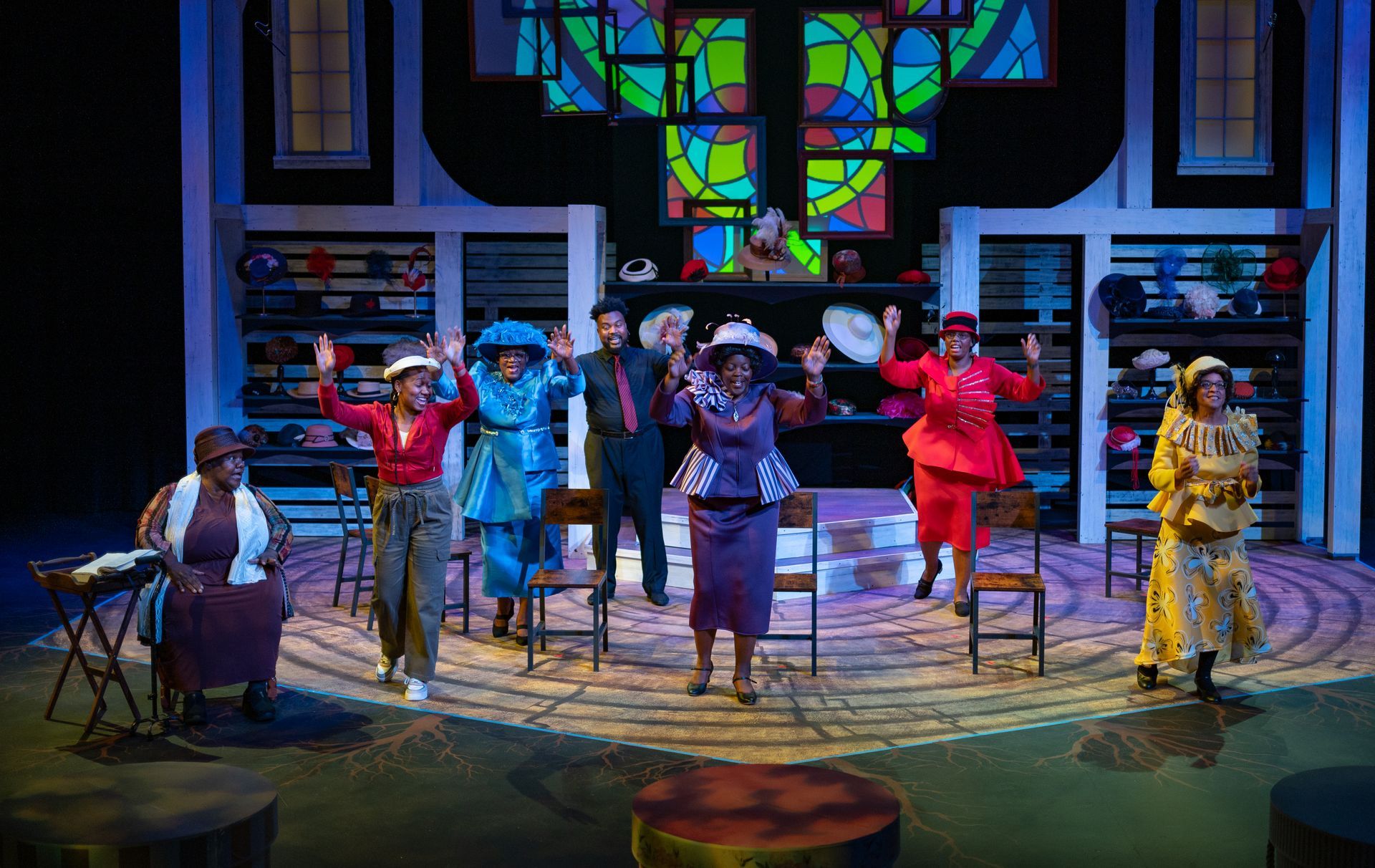 A group of people in hats dance on a stage. Bright colors, stained glass, and a hat shop background.