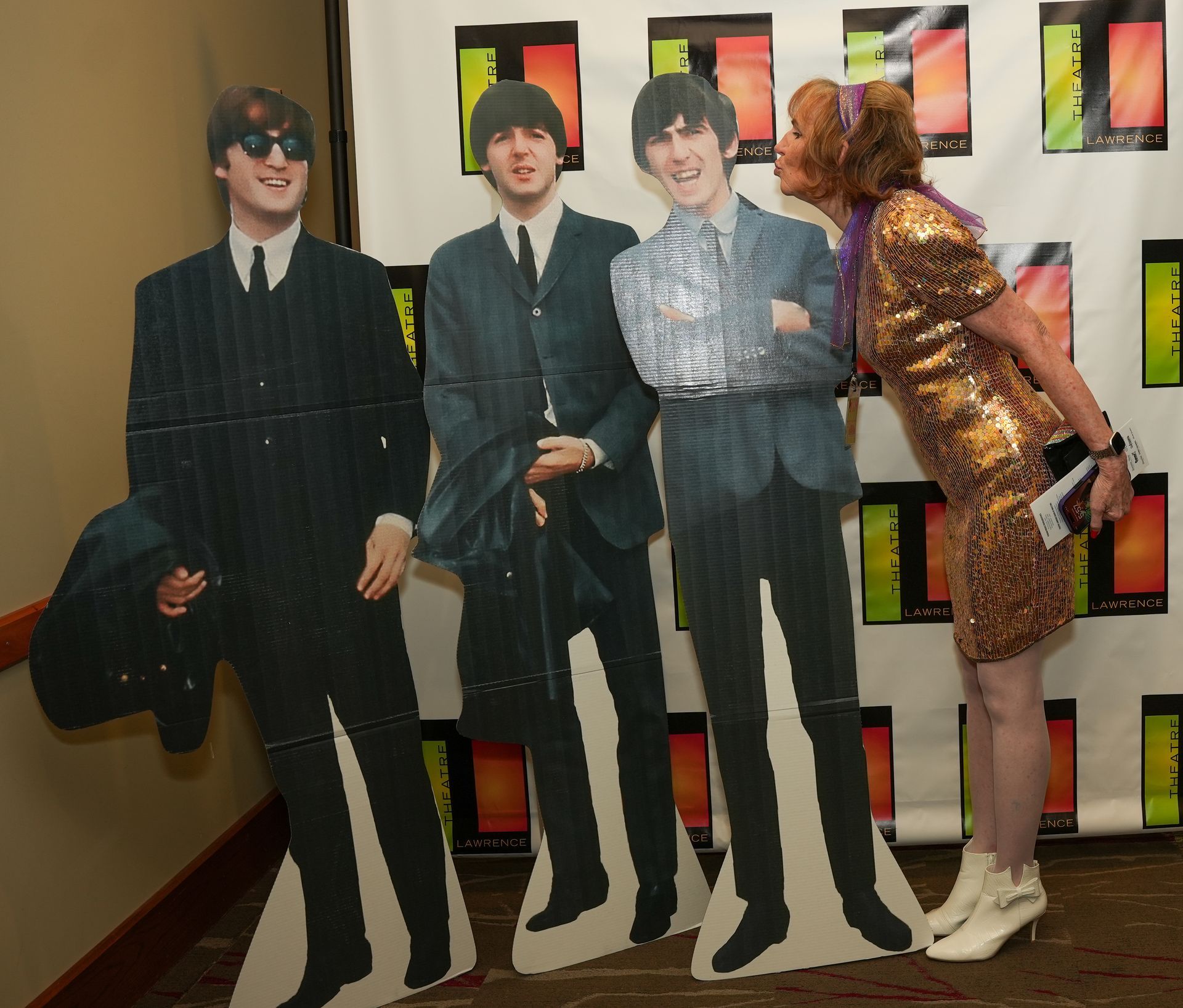 Woman in gold dress kissing cardboard cutout of a Beatle. Other cutouts of Beatles are present.
