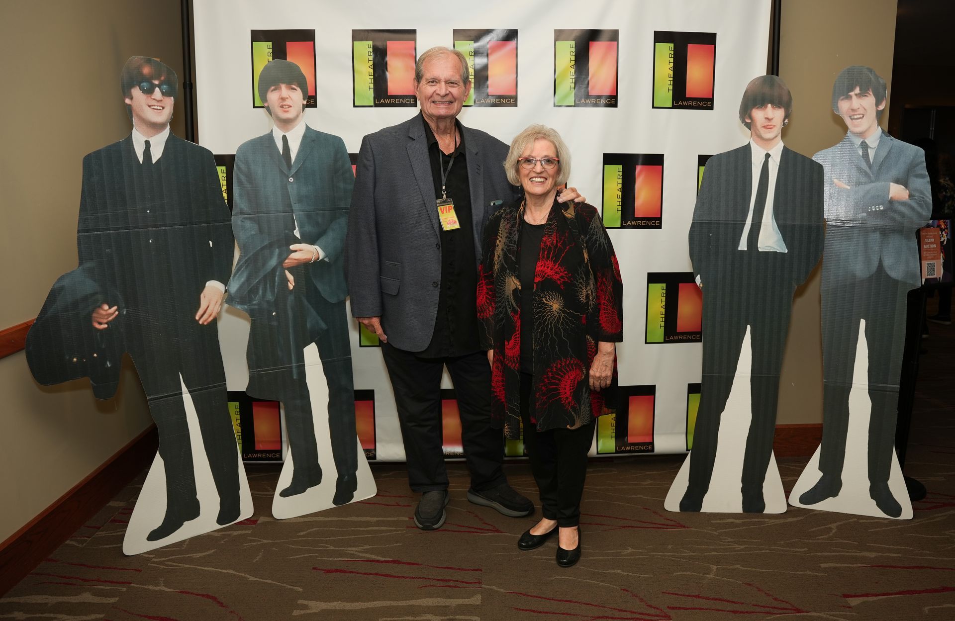 Couple poses with Beatles cutouts at an event. White backdrop with colorful rectangles.