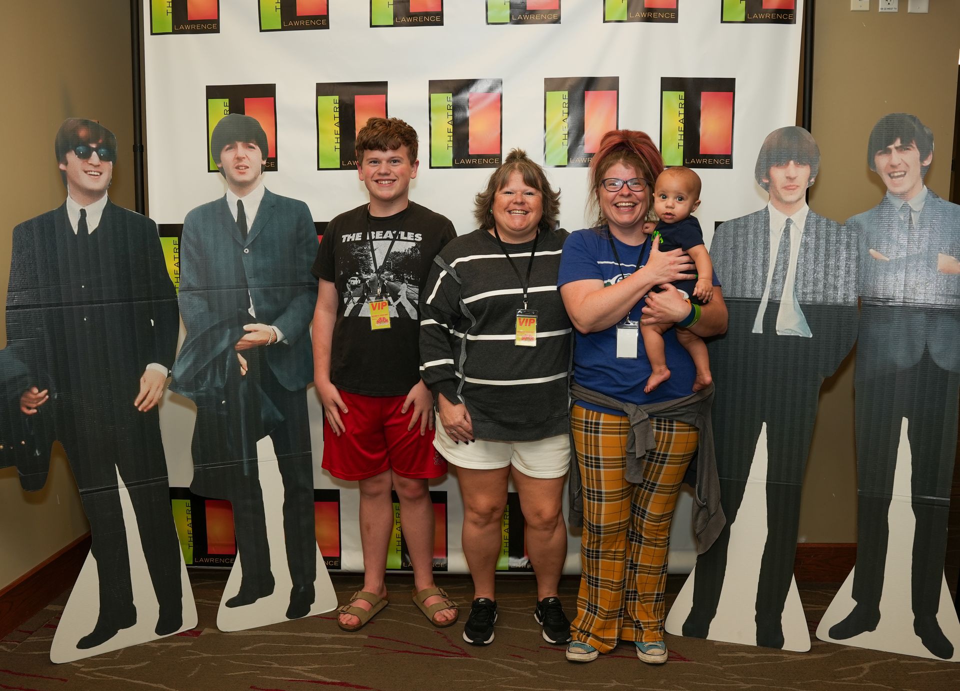 People pose with Beatles cutouts at an event. Three adults and a baby smile, banner background.