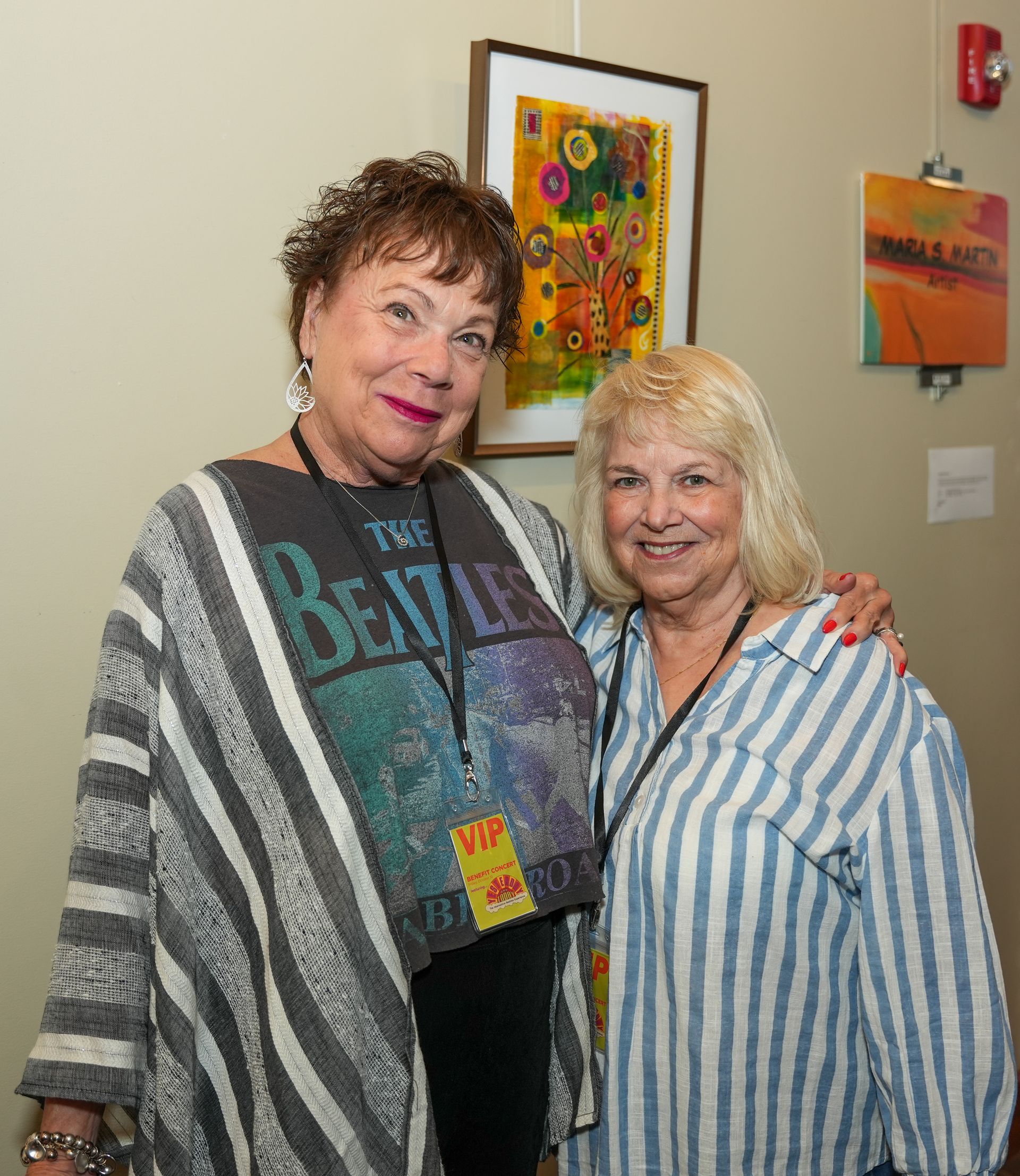 Two women smiling, posing at an art gallery. The woman on the left is wearing a Beatles shirt and a striped shawl.