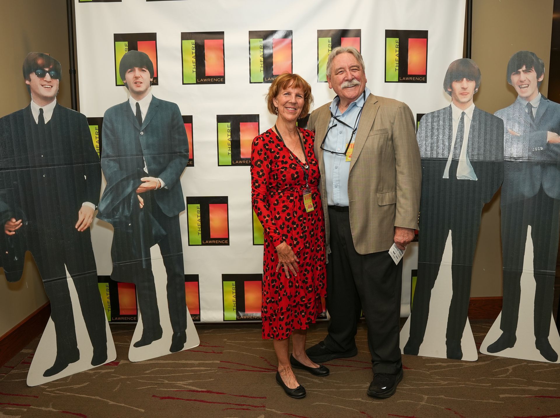 Couple poses with Beatles cutouts at an event, red dress, tan blazer, backdrop with logos.