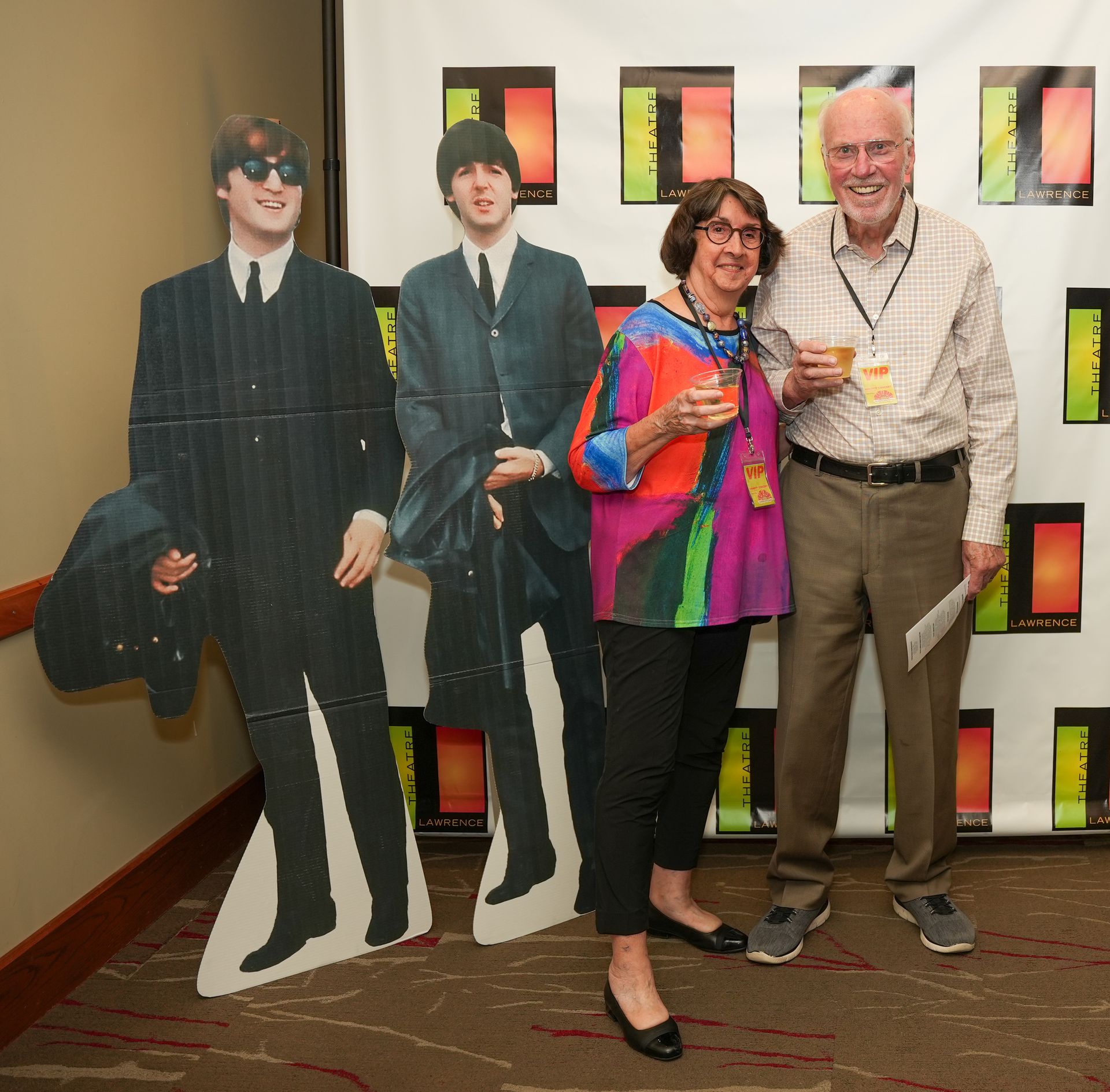 Two people pose with Beatles cutouts. Woman in colorful shirt and man in plaid shirt hold drinks.