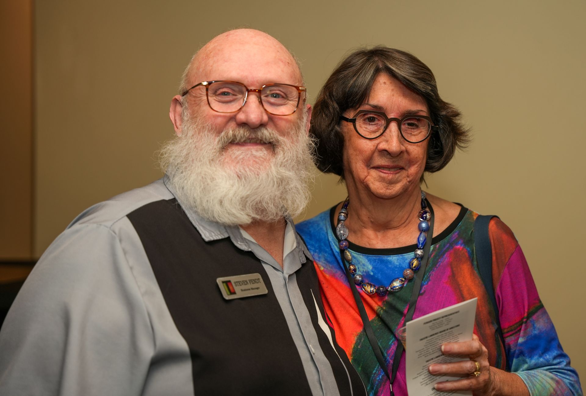 Man with a white beard and woman with glasses pose together. Man wears a shirt with a name tag.