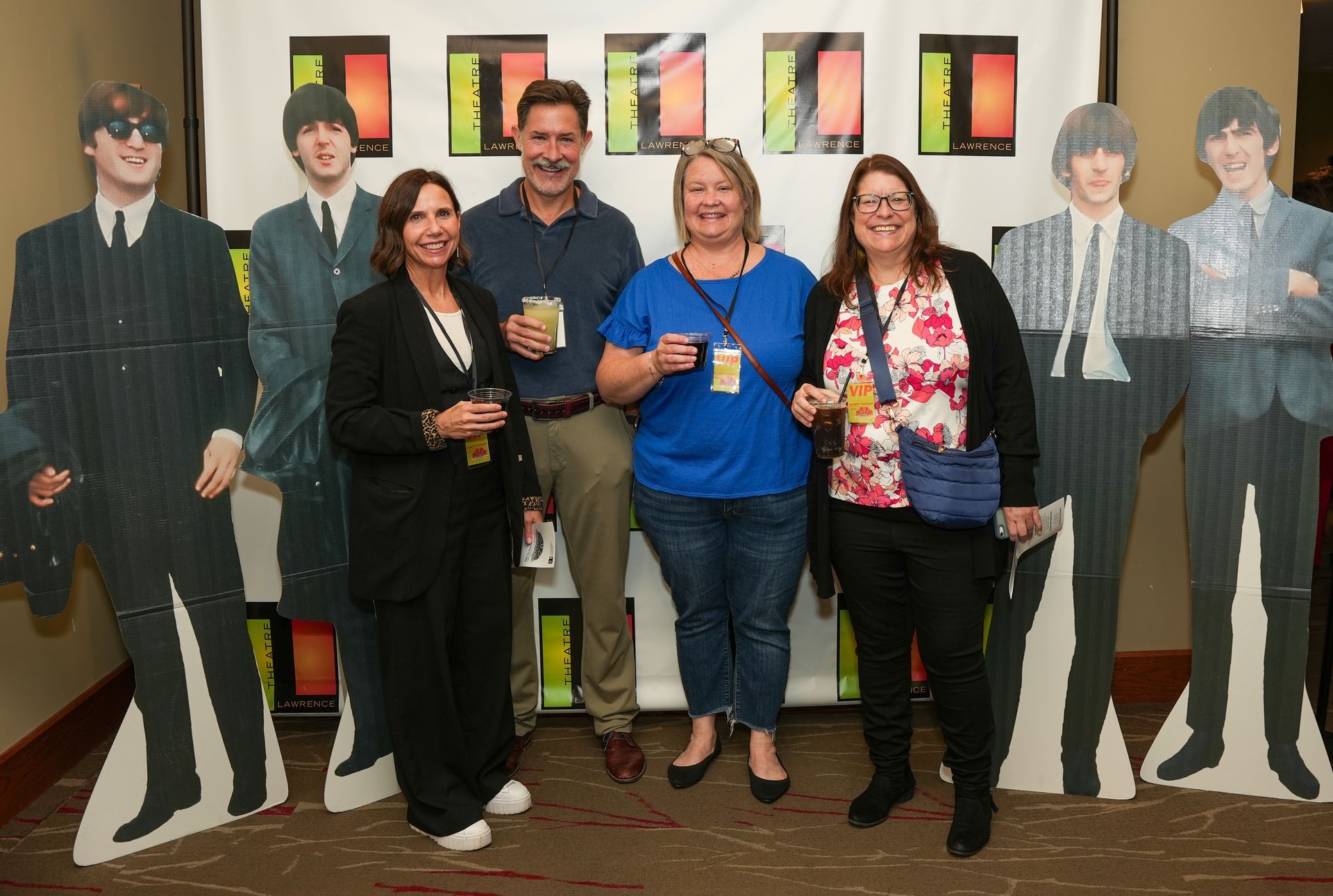 Group of people pose with Beatles cutouts at an event, holding drinks, smiling.