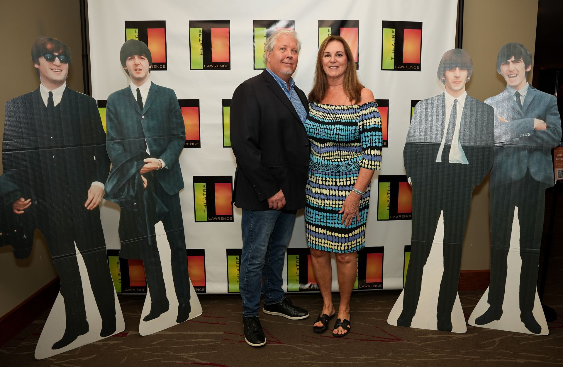 A couple poses with Beatles standees at an event. They stand in front of a backdrop with colorful squares.