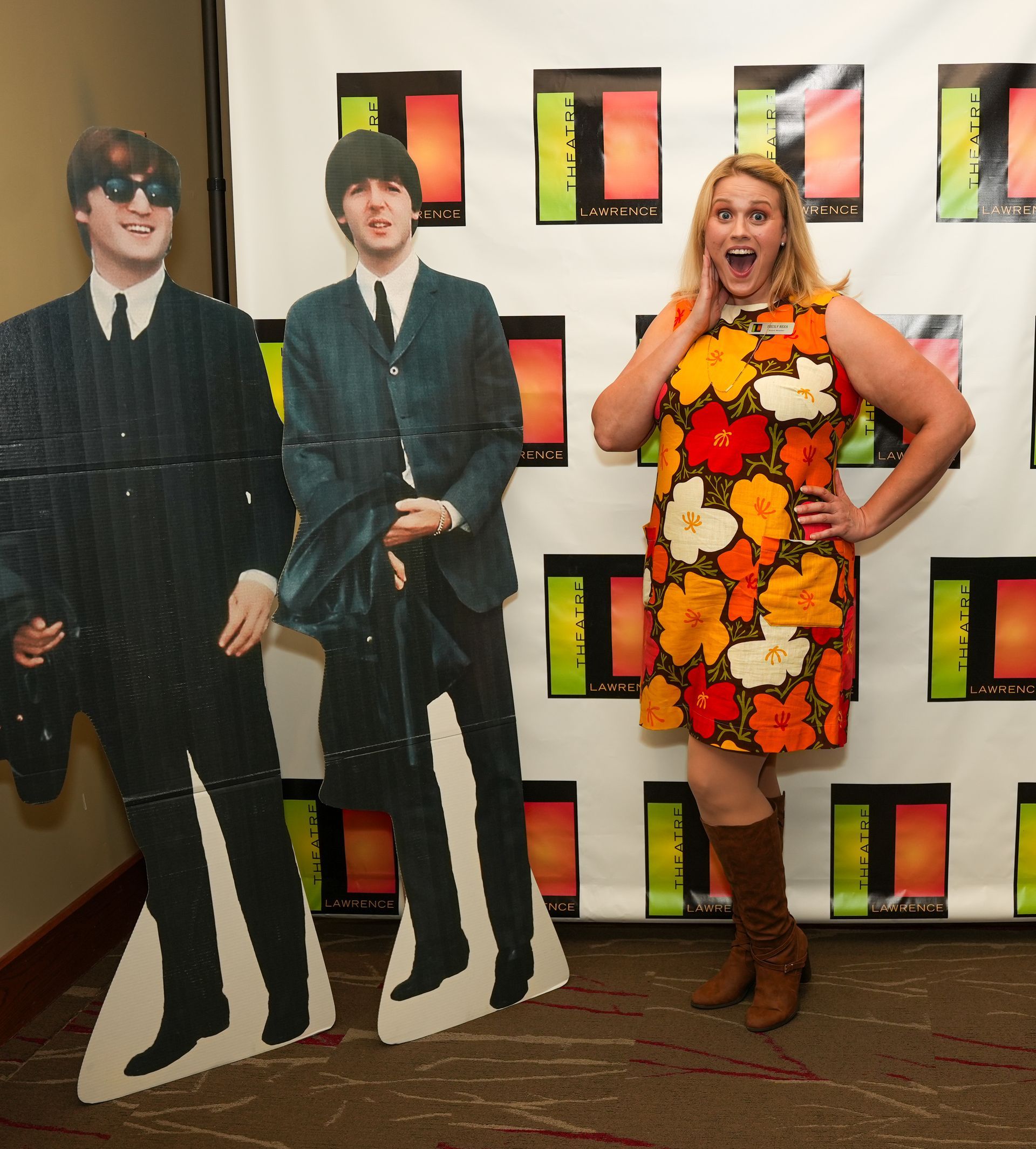 Woman in floral dress, poses with Beatles cutout, surprised expression, boots, event backdrop.