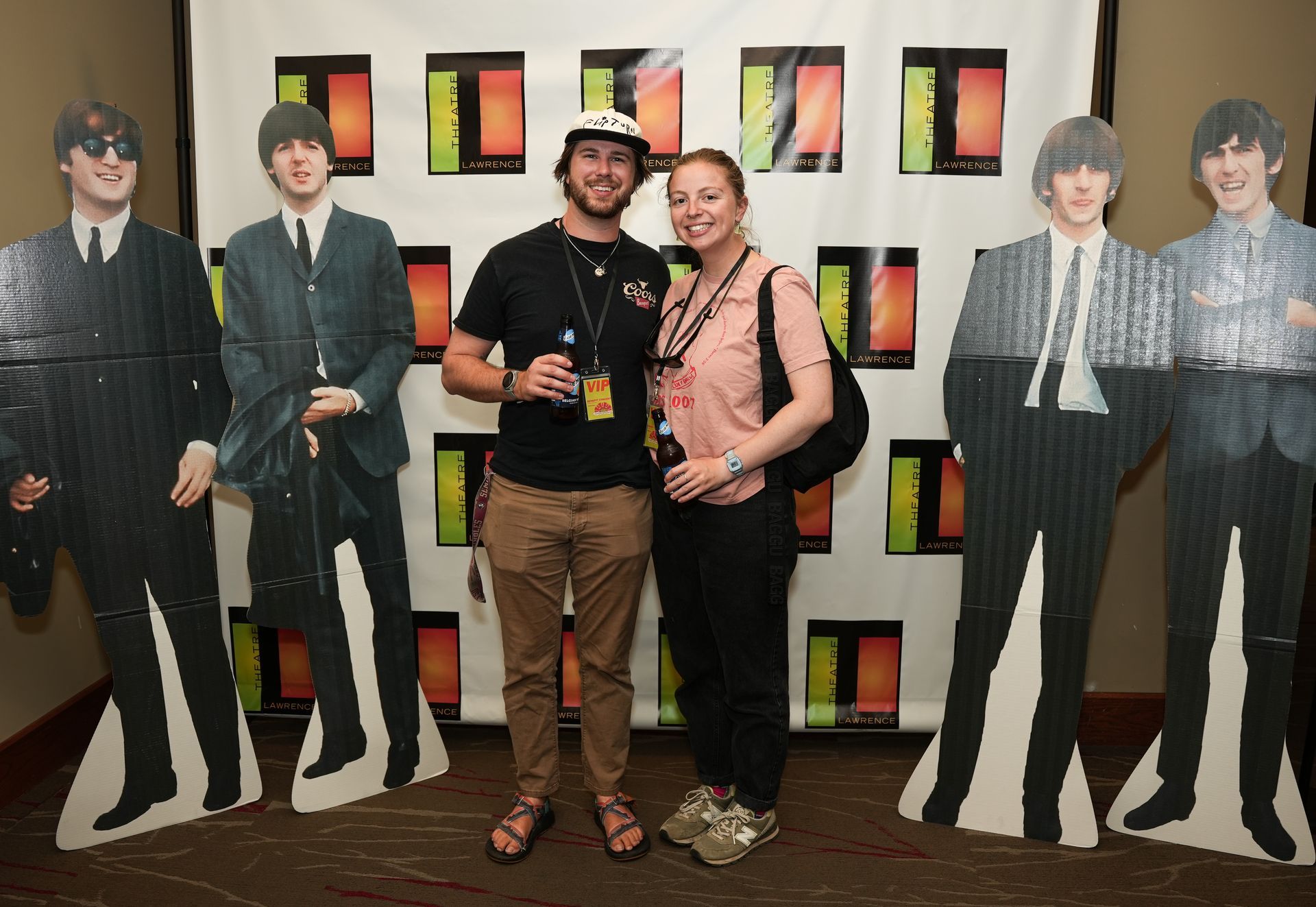 Two people pose with Beatles cutouts in front of a branded backdrop.