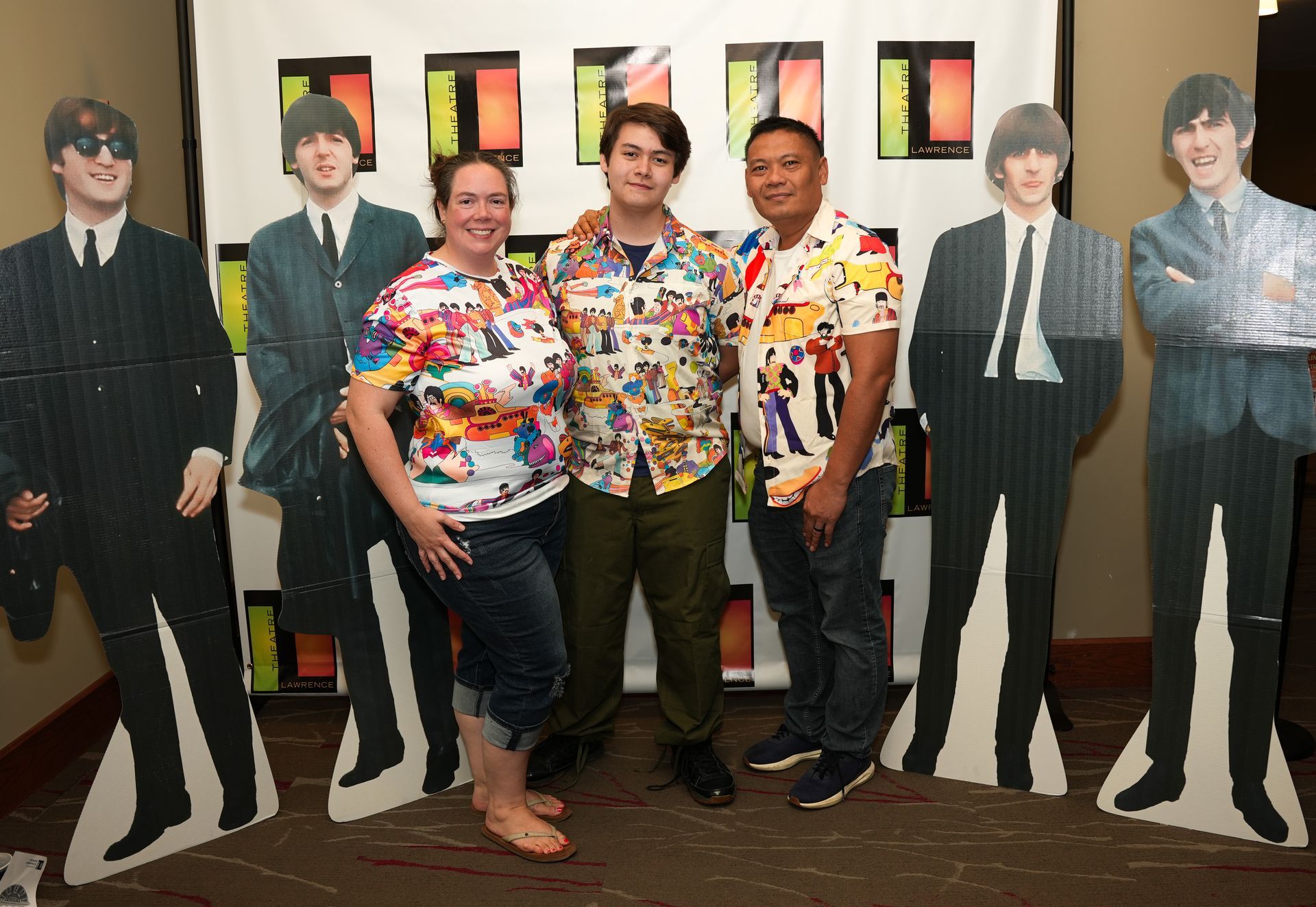 Three people pose with Beatles cardboard cutouts at an event. They wear Beatles-themed shirts.