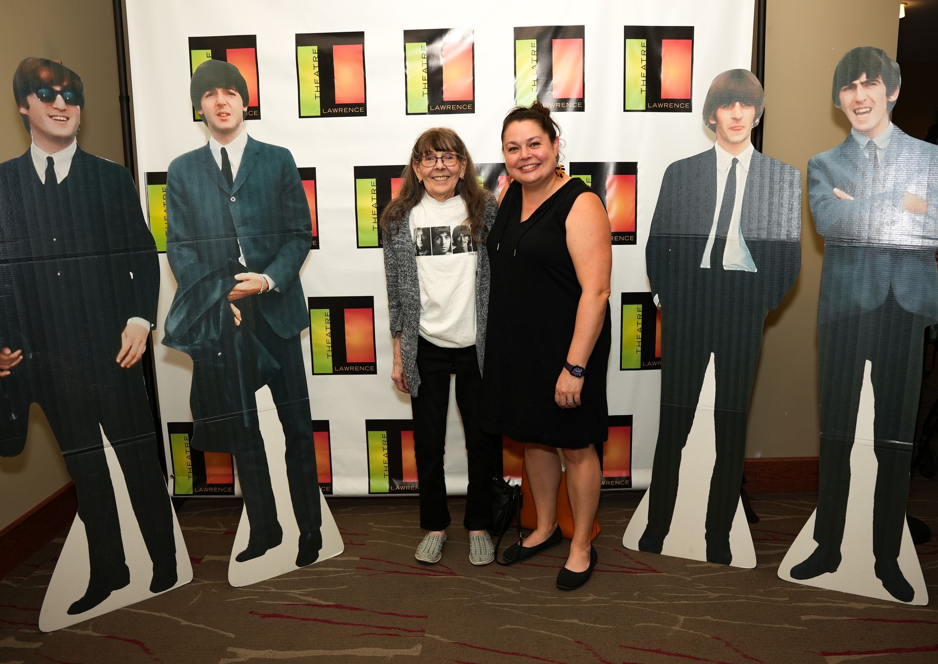 Two people pose with Beatles cardboard cutouts at an event. They are in front of a backdrop with colorful squares.