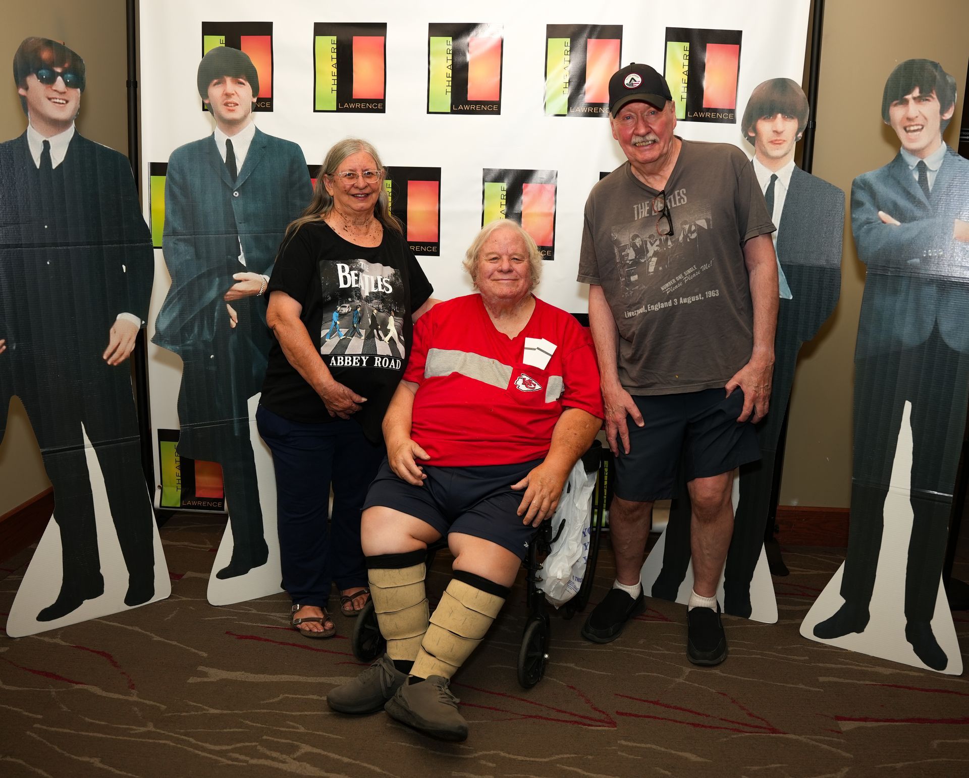 Three people pose with Beatles standees; one in a wheelchair, all smiling at a convention.
