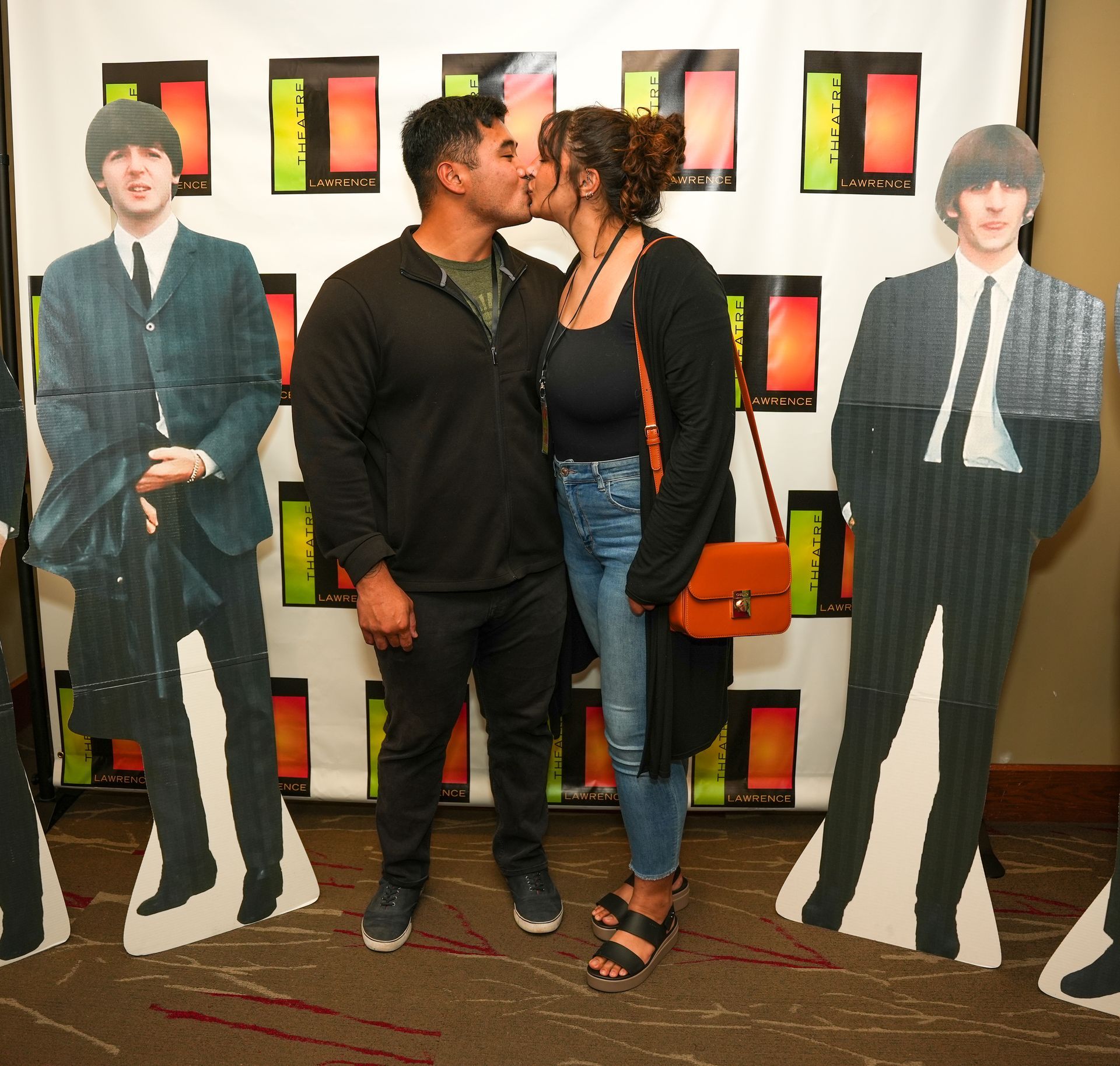 Couple kissing between cardboard cutouts of Beatles members. They stand before a backdrop with colored rectangles.