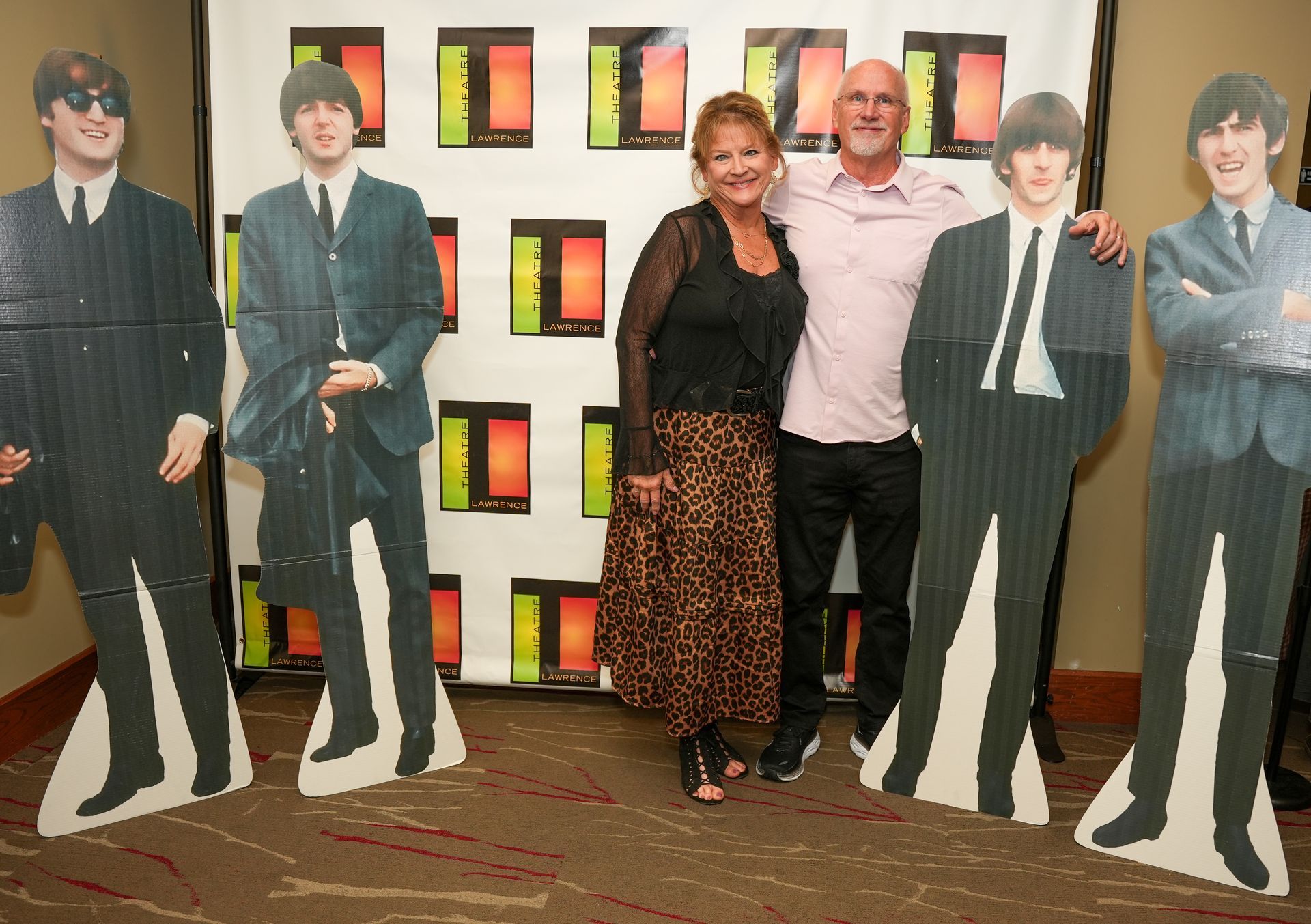 Two people pose with Beatles cutouts in front of a backdrop. Woman in leopard skirt, man in pink shirt.