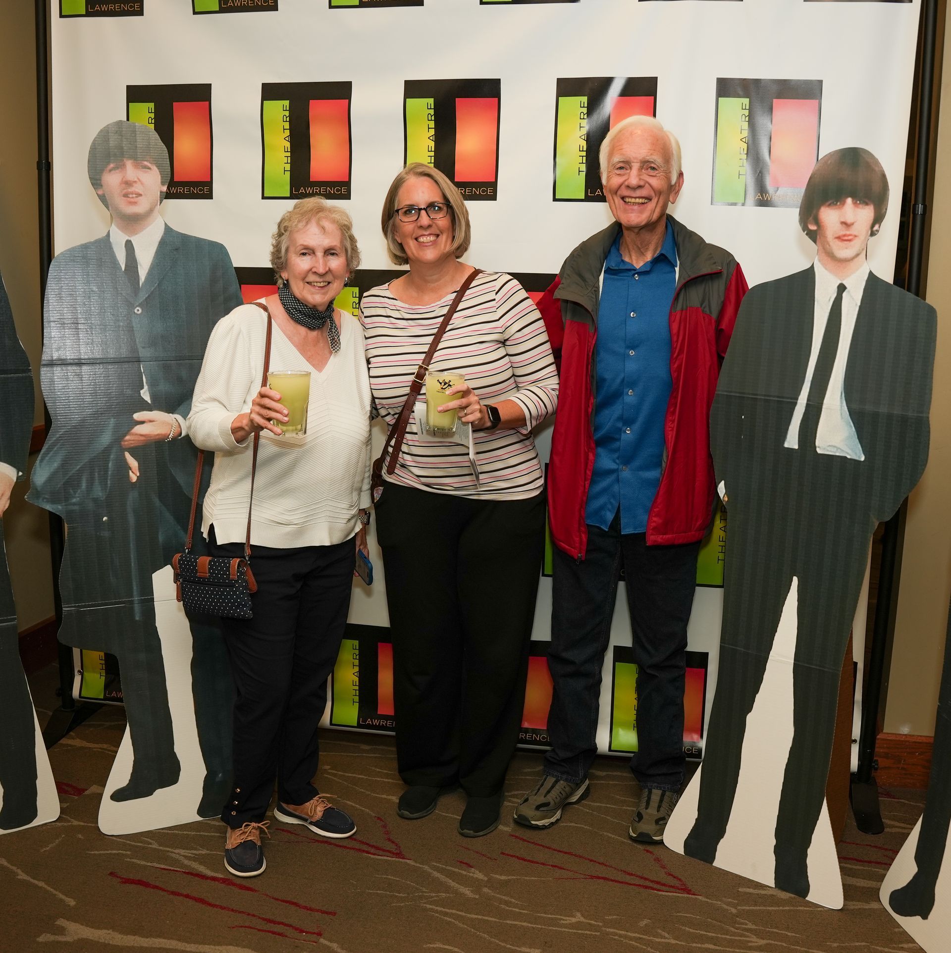 Three people smiling, posing with Beatles cutouts. They are holding drinks, in front of a backdrop.