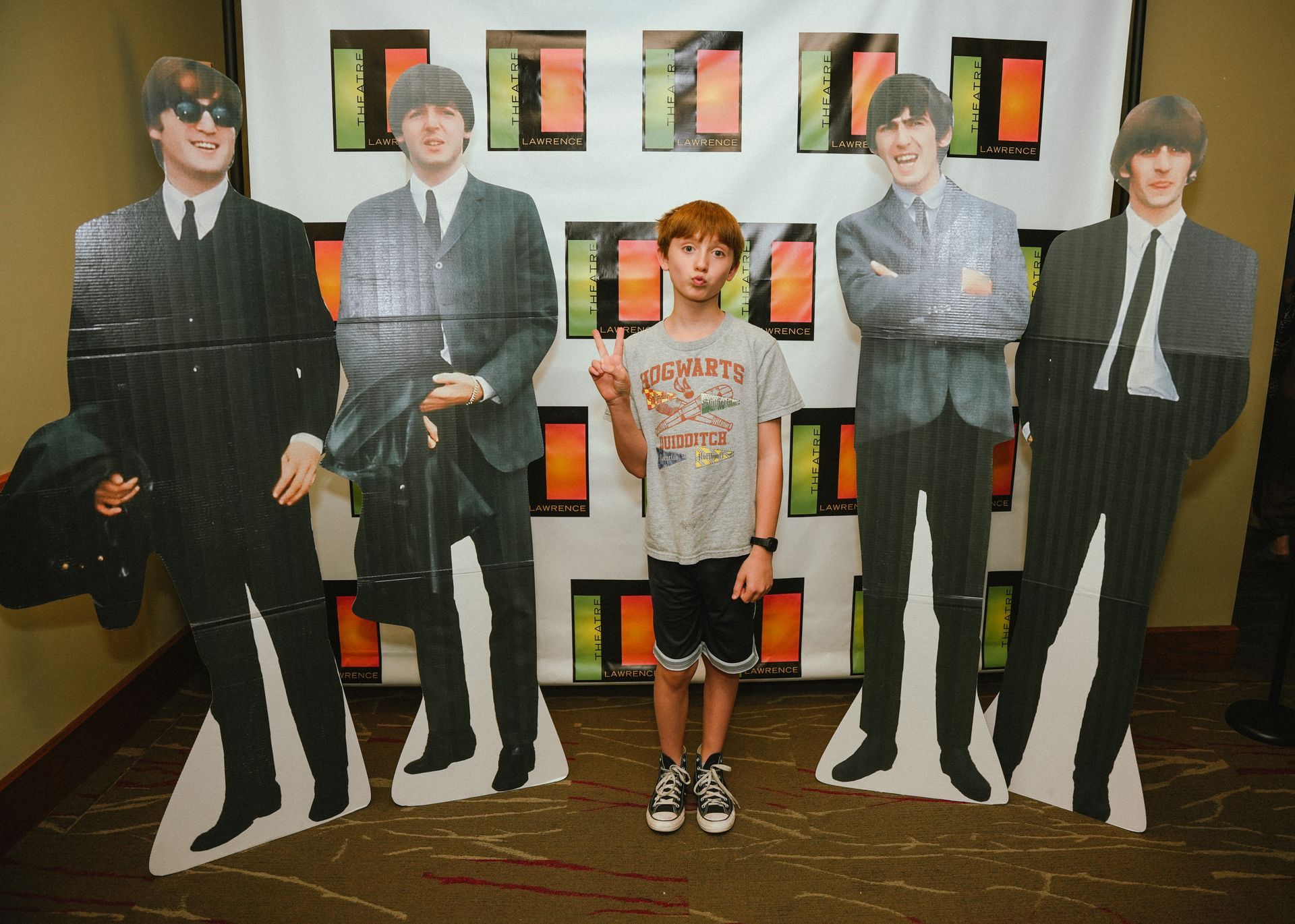 Boy posing with cardboard cutouts of The Beatles. He gives the peace sign. Background: colorful squares.