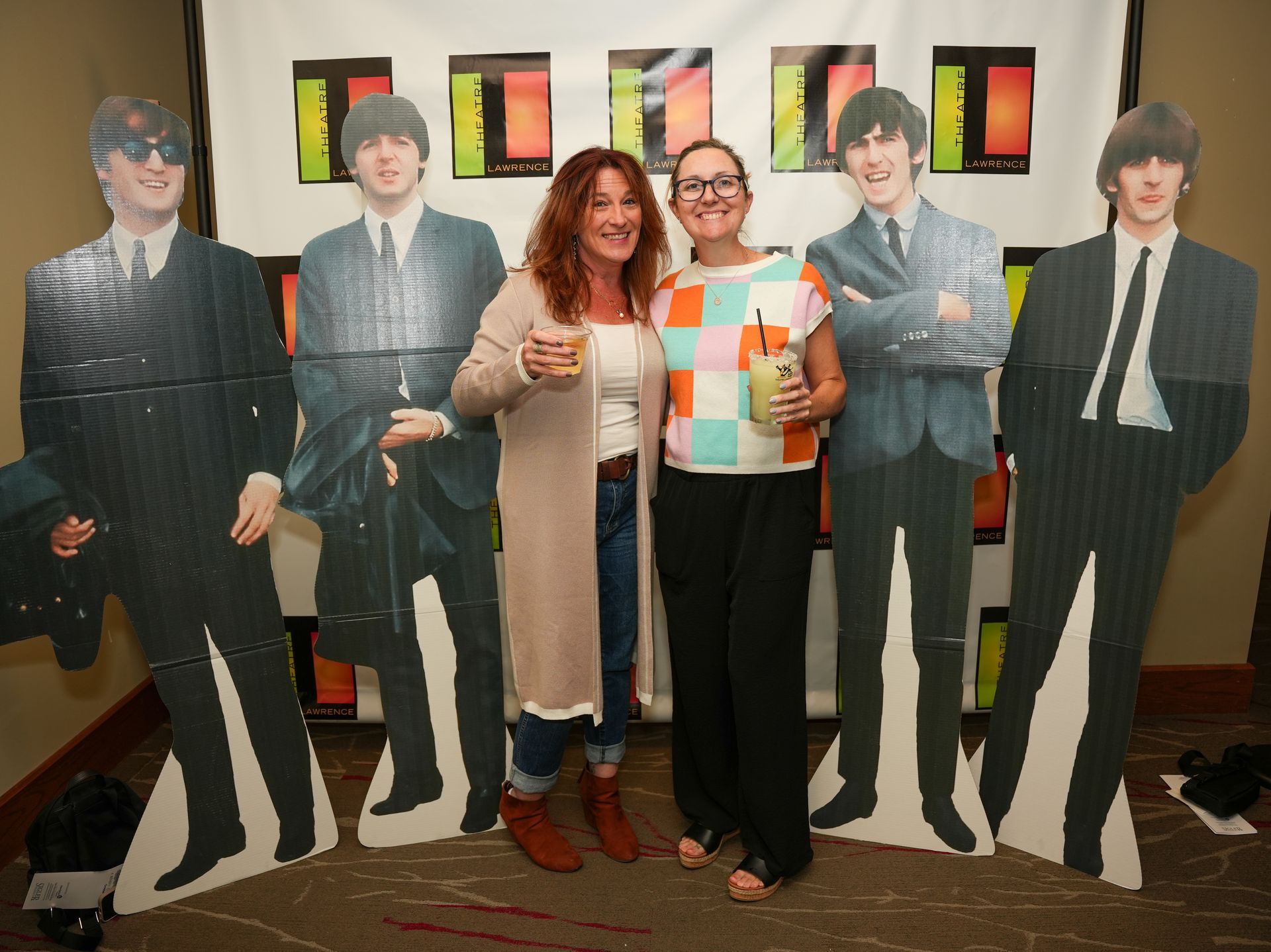 Two women pose with Beatles cutouts; one holds a drink, smiling in front of a colorful backdrop.