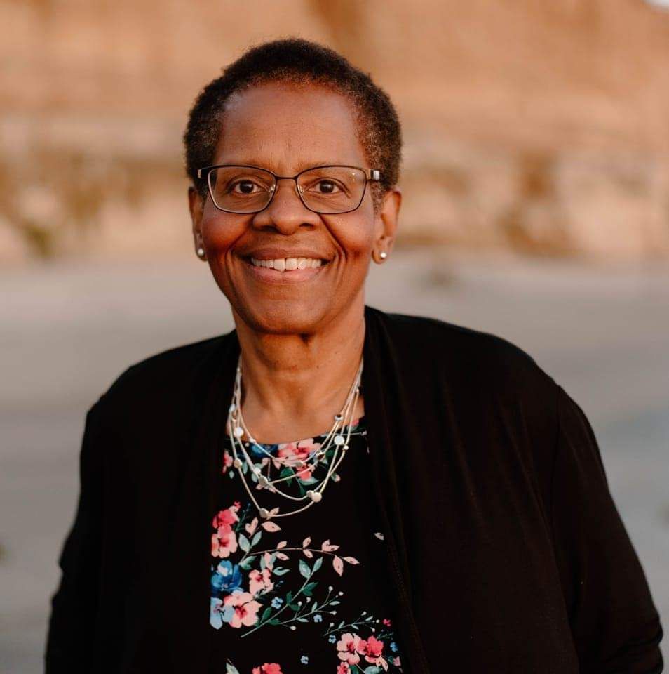 Smiling Black woman in glasses, floral top, and black jacket, outdoors at sunset.