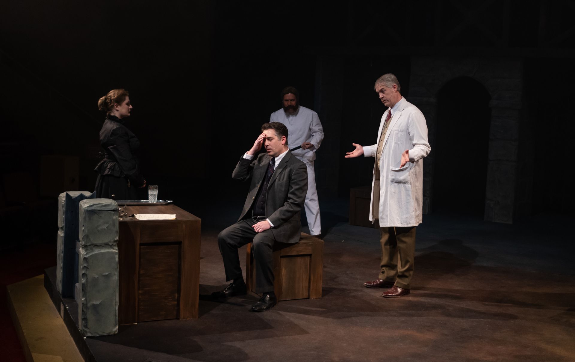 Four people on stage. A man in a suit sits, distressed. A doctor gestures. Another man and woman stand by. 