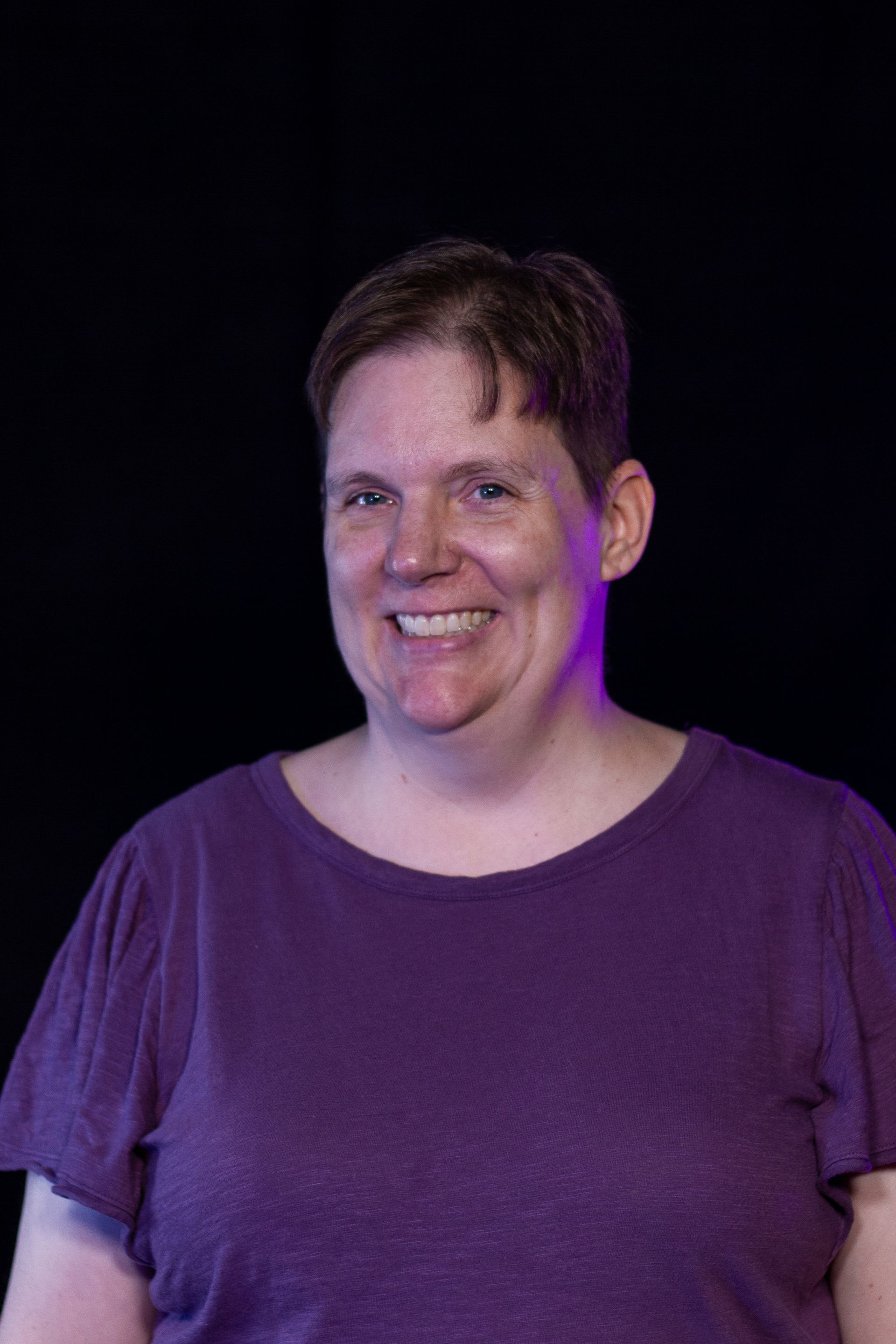 A woman in a purple shirt is smiling in front of a black background.