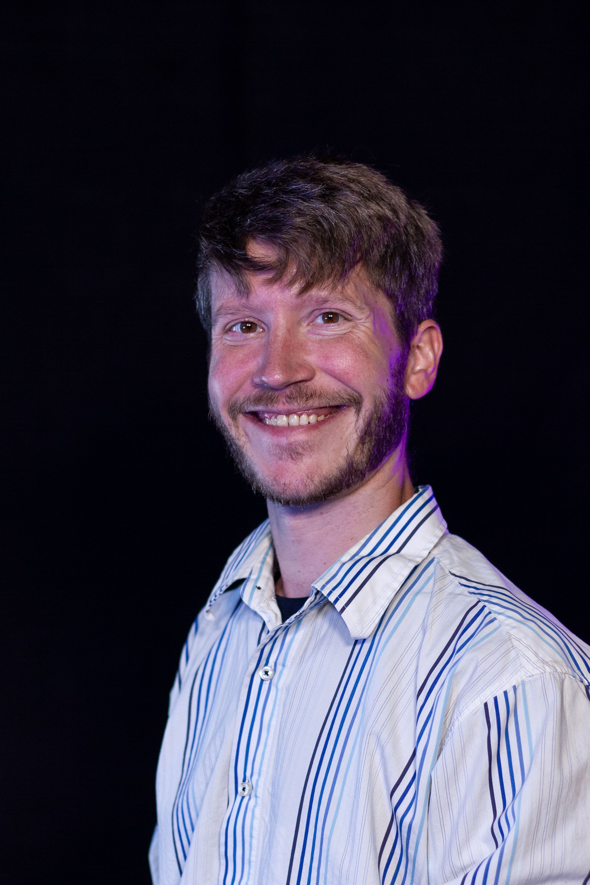 Smiling man with a beard in a striped shirt, posed against a black background.
