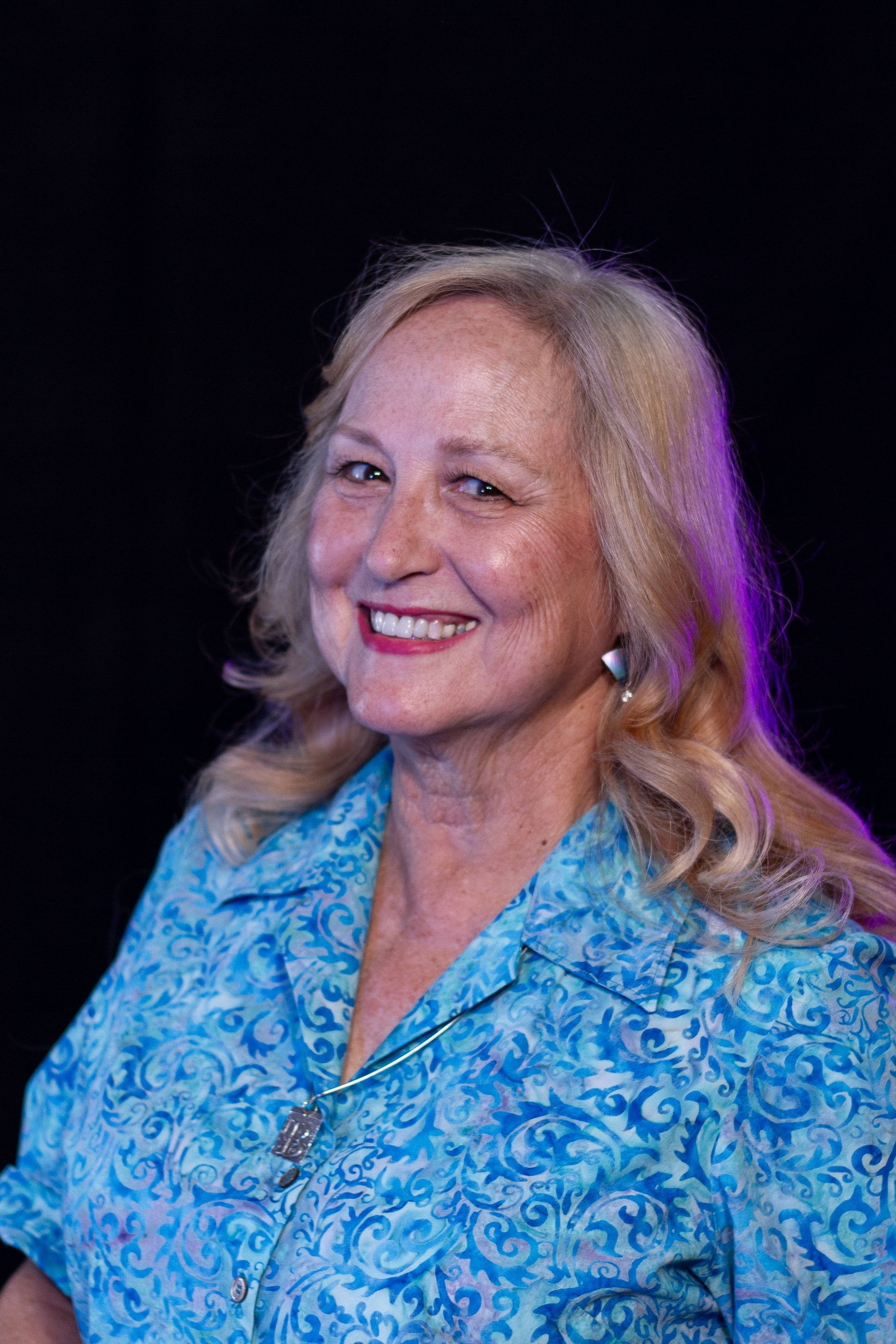 Woman with blonde hair smiles, wearing blue patterned top, black background.