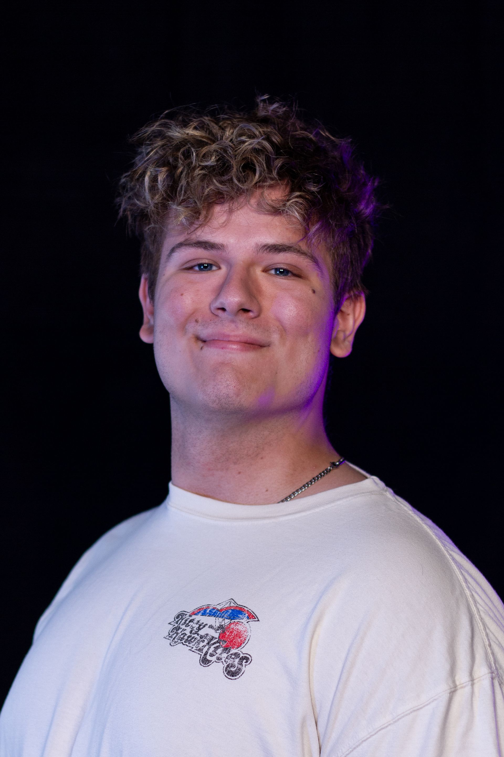 Young man with curly hair smiles, wearing a cream shirt with floral design; black background.