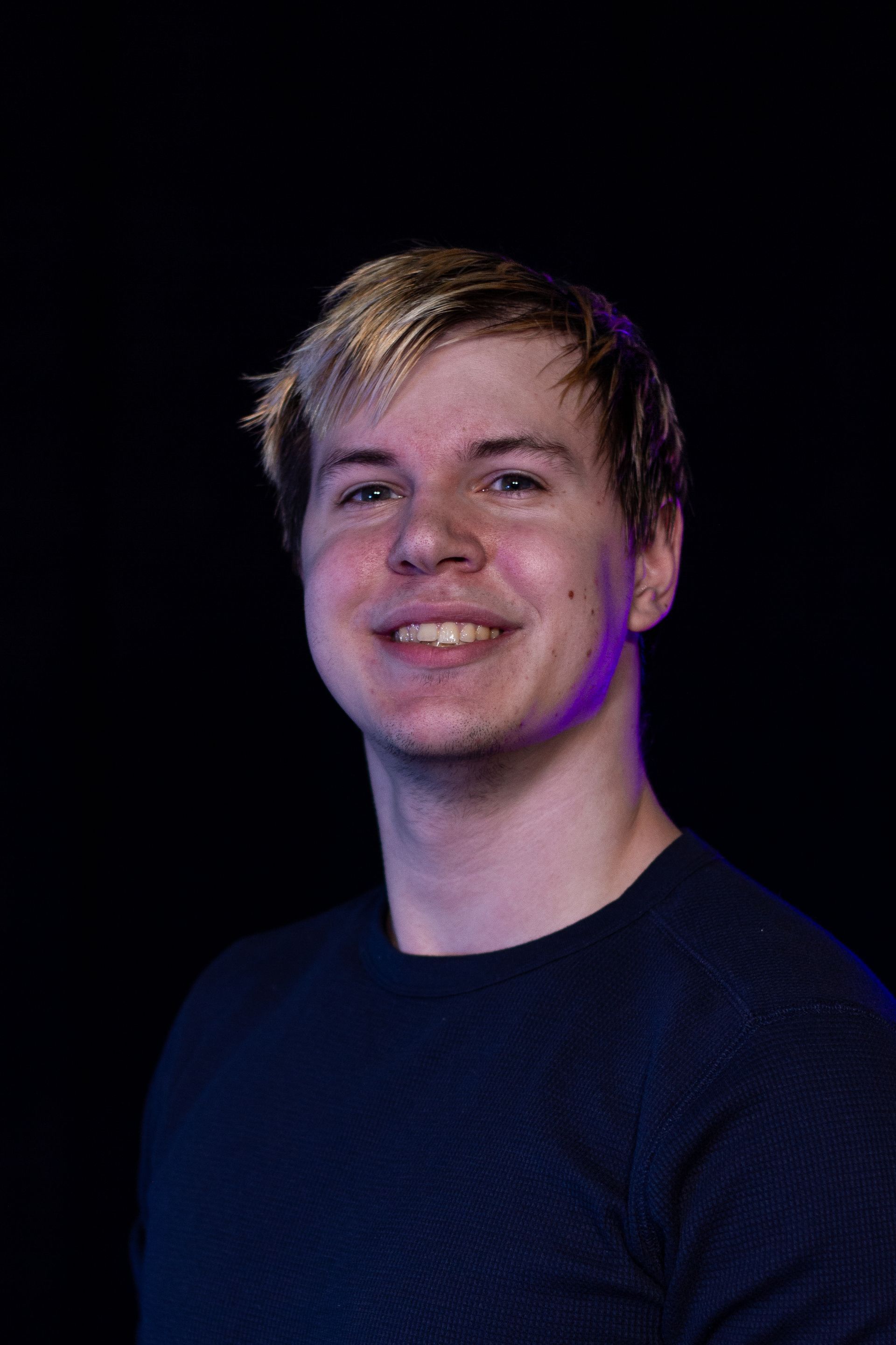Young man with blond hair smiles, wearing a blue sweater against a black background.