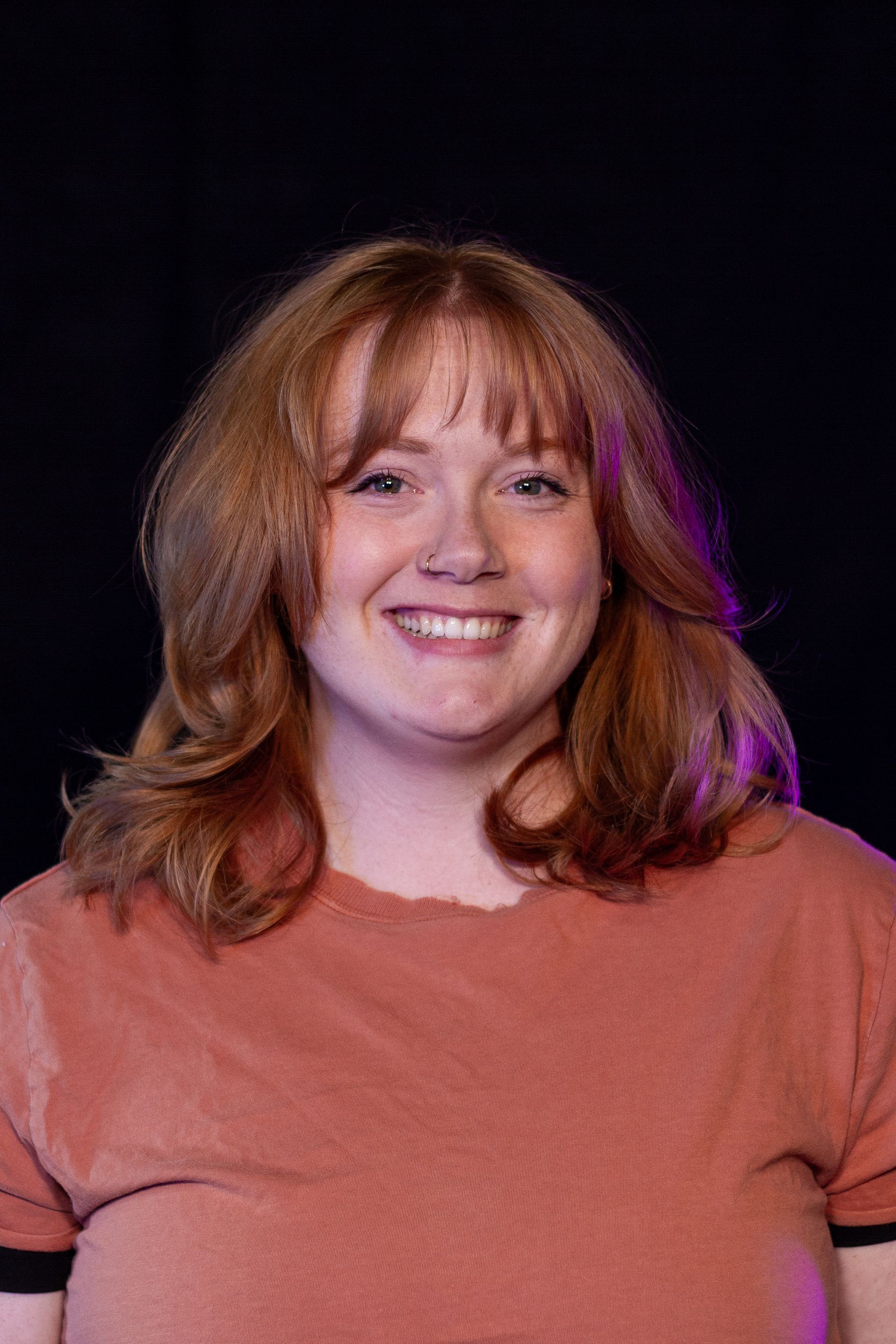 Woman with red hair smiling, wearing a rust-colored shirt. Black background.
