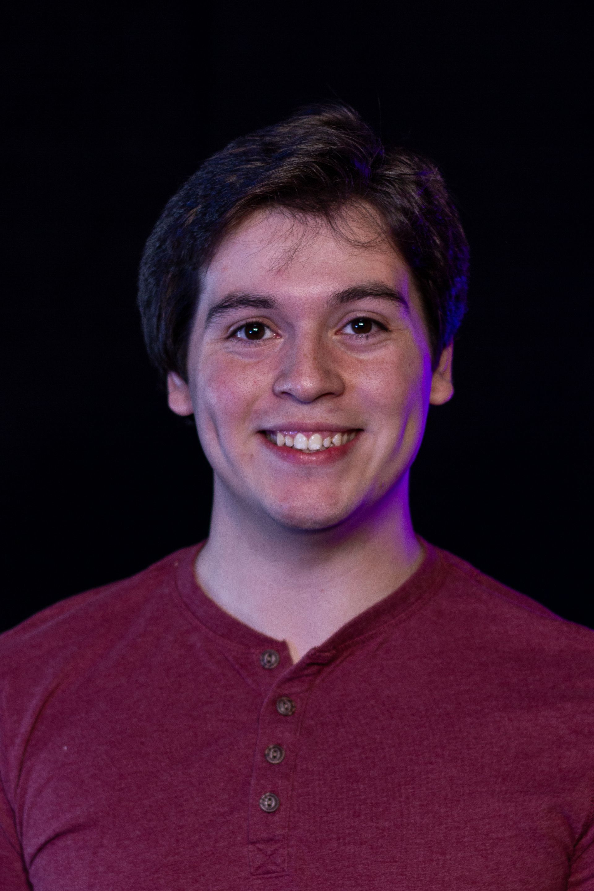 Young man with a warm smile, wearing a maroon shirt against a black background.