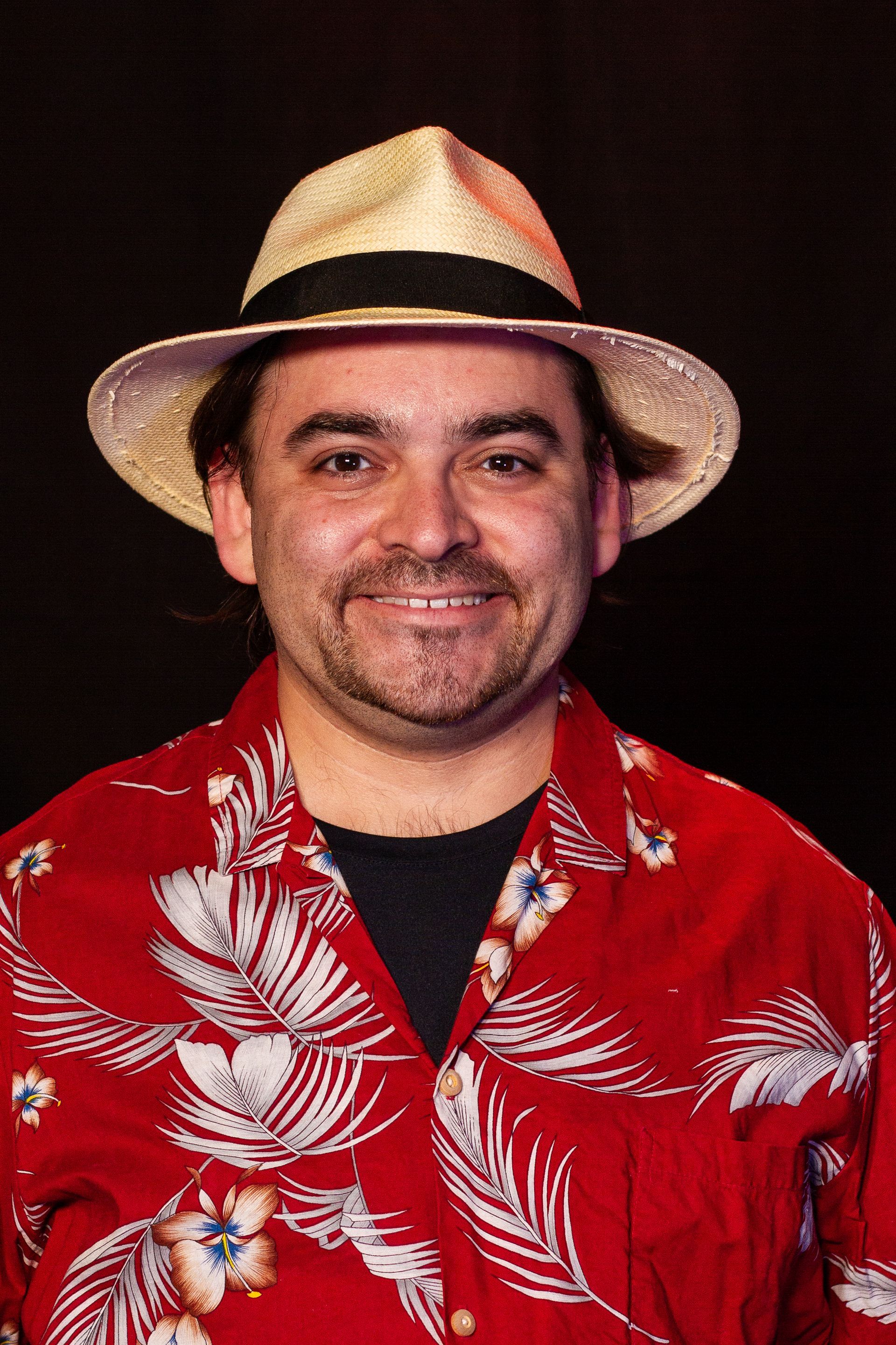 Man in red Hawaiian shirt and straw hat smiling; black background.