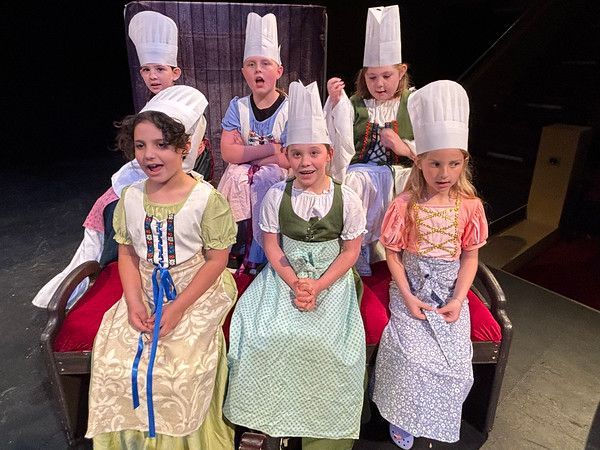 Children in chef hats and aprons on a stage. They are singing, smiling, and sitting on a bench.